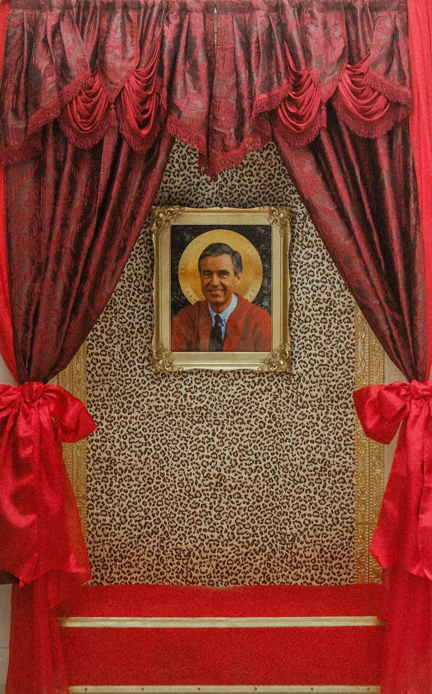 A decorative display featuring a framed portrait of a smiling man in a judicial robe, set against a leopard print background surrounded by red curtains with tassels. The cushion beneath the portrait matches the leopard print background.