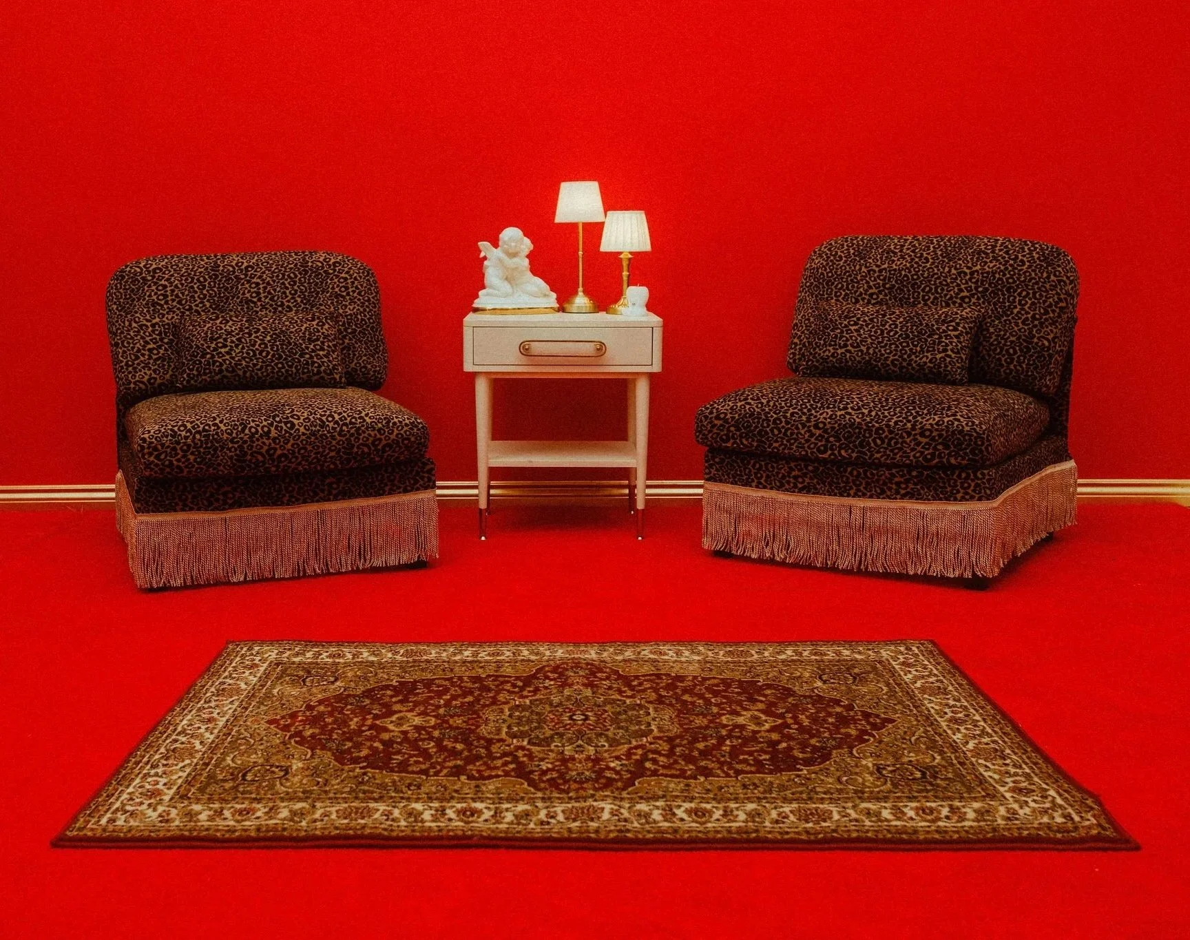 Two leopard print armchairs with a matching footstool on a red carpet, a white side table with a white figurine, two white lamps, and vase, and a Persian-style rug in front of them against a red wall.