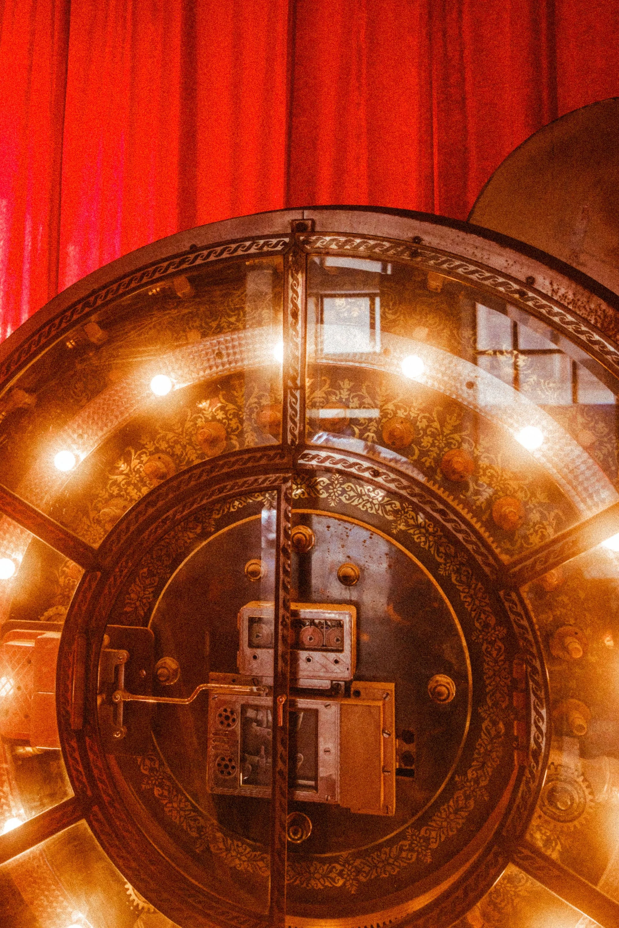 Inside view of an ornate, vintage, circular coin bank door, featuring intricate gold patterns and a lock mechanism in the center, with red curtains in the background.