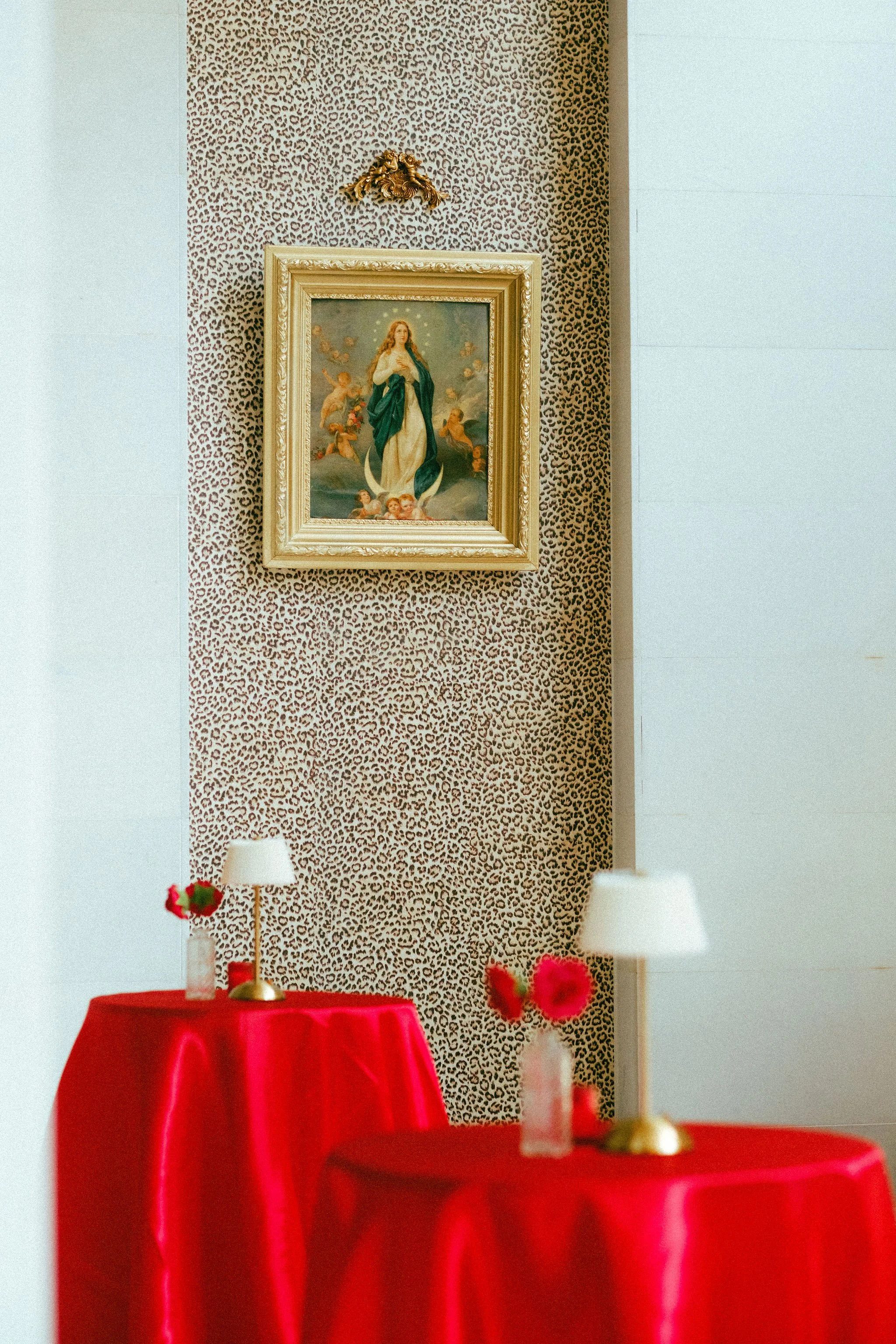 Interior room decorated for a celebration with a red tablecloth, white lamps, vases with red flowers, a leopard print wallpaper, and a framed religious painting of the Virgin Mary with angels.