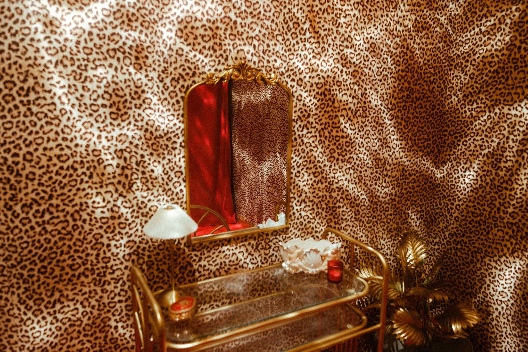 A room with leopard print wallpaper, a gold-framed mirror, a gold cleaning cart with a glass surface, and decorative plants.
