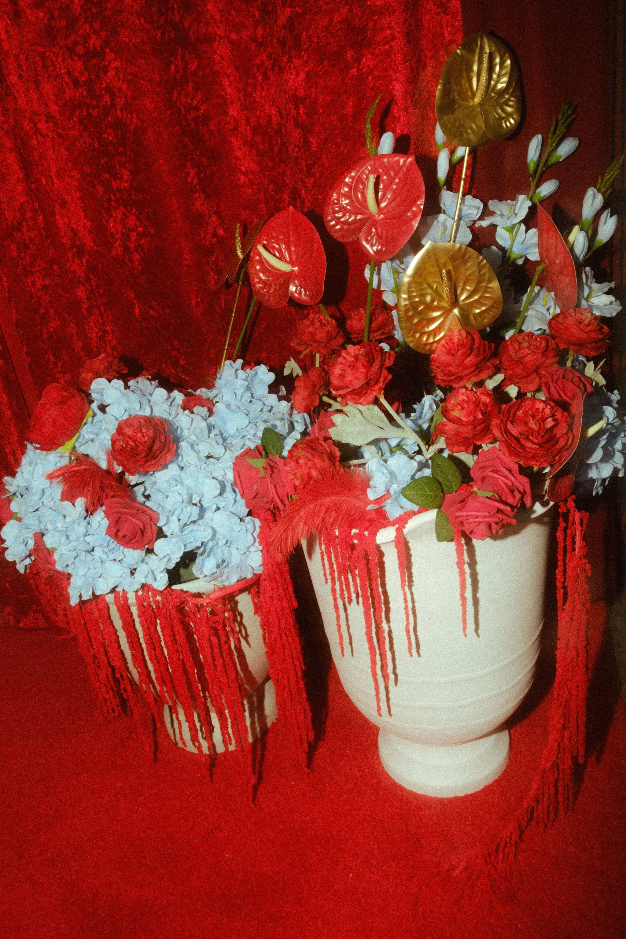 Two flower arrangements in tall white pots against a red background. The arrangements include red roses, white flowers, red anthuriums with yellow centers, and some greenery.