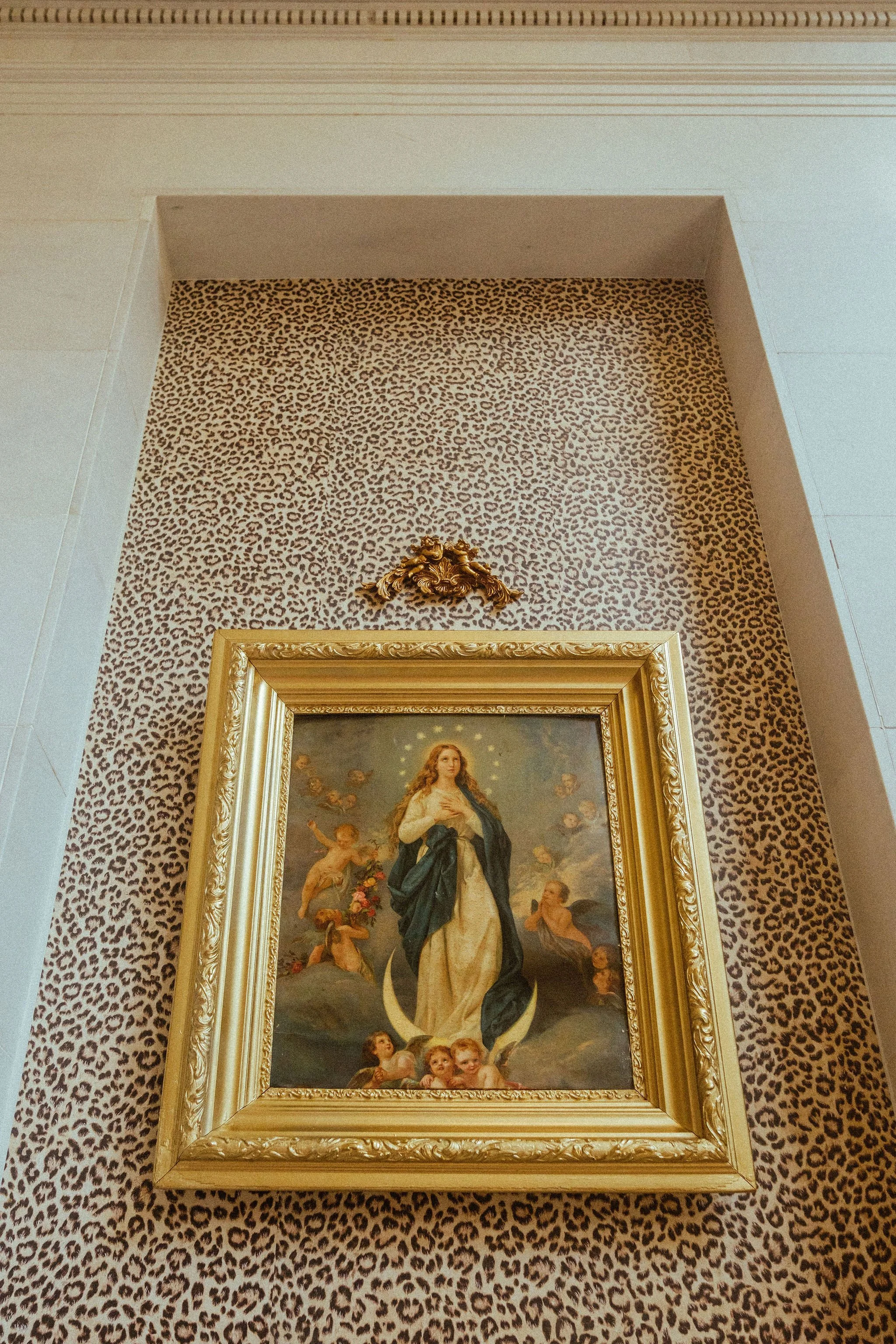 A framed religious painting of the Virgin Mary surrounded by angels, set against a leopard-print wallpaper background, with intricate gold accents above and around the frame.