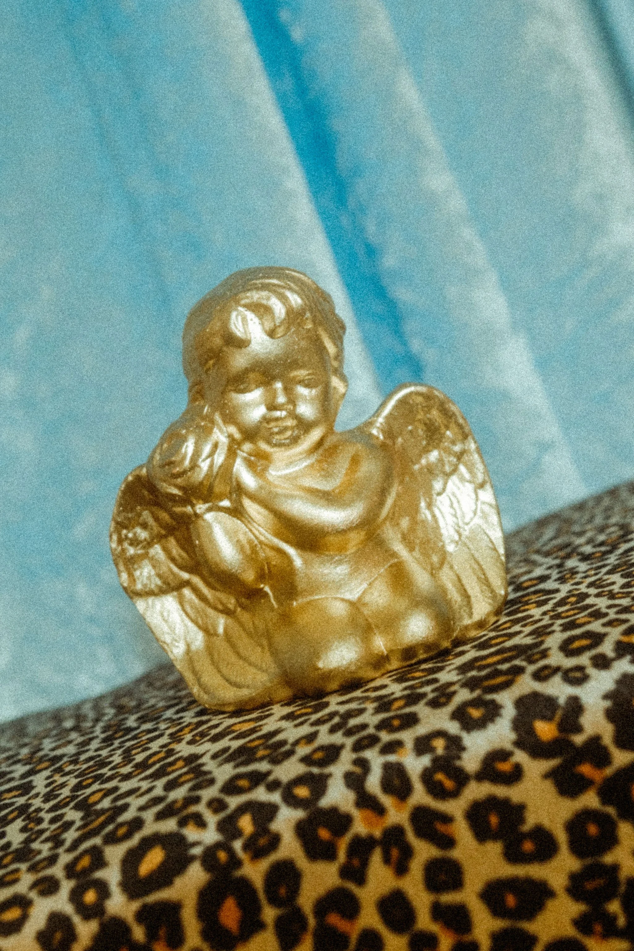 Gold-colored angel figurine with closed eyes, smiling face, and hands on cheeks, placed on leopard print fabric with a blue background.