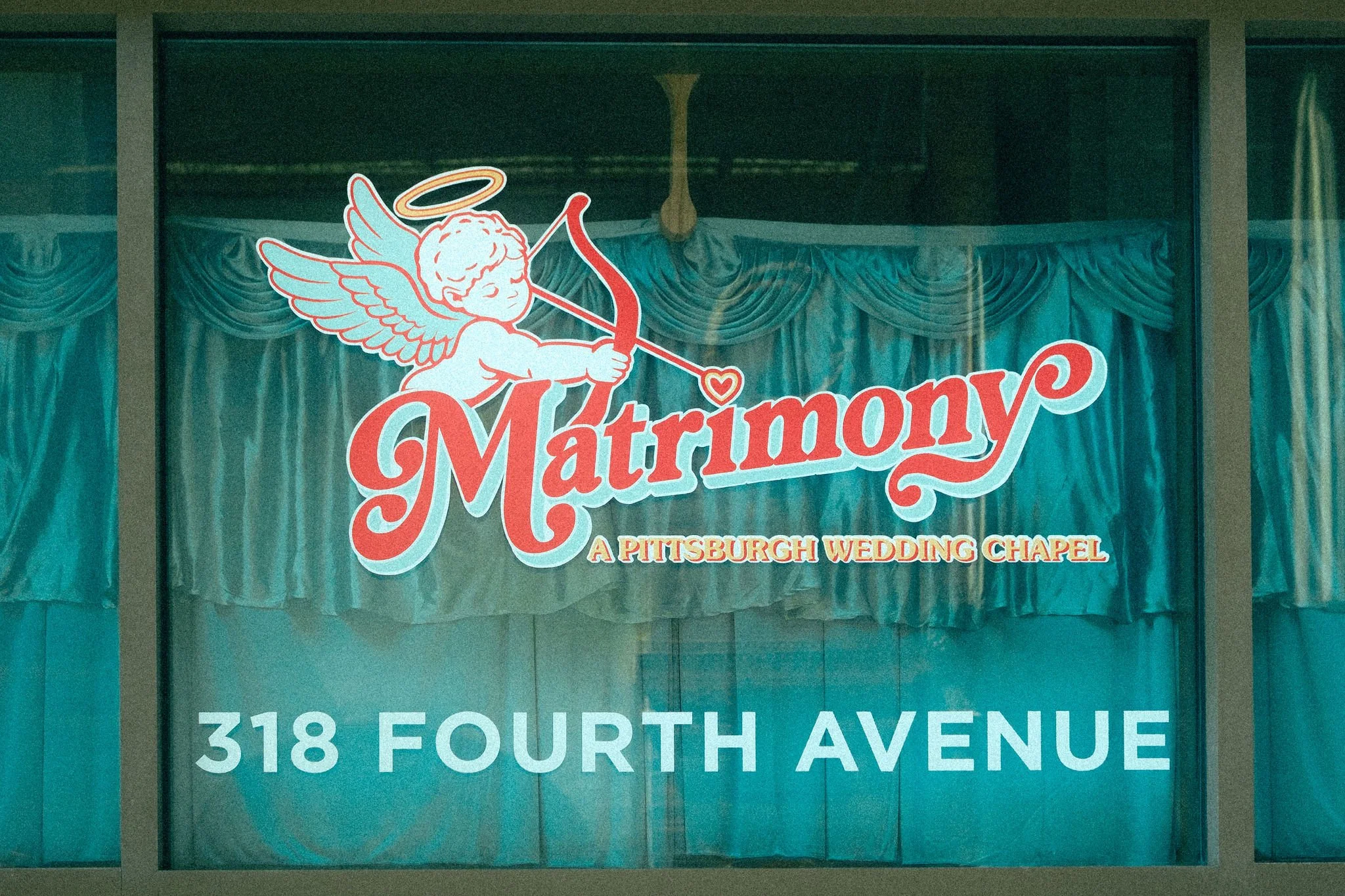 Sign for a wedding chapel with a logo of a cherub with wings and a halo, holding a bow and arrow, and the word 'Matrimony' in red cursive lettering. The sign also lists a street address, 318 Fourth Avenue.