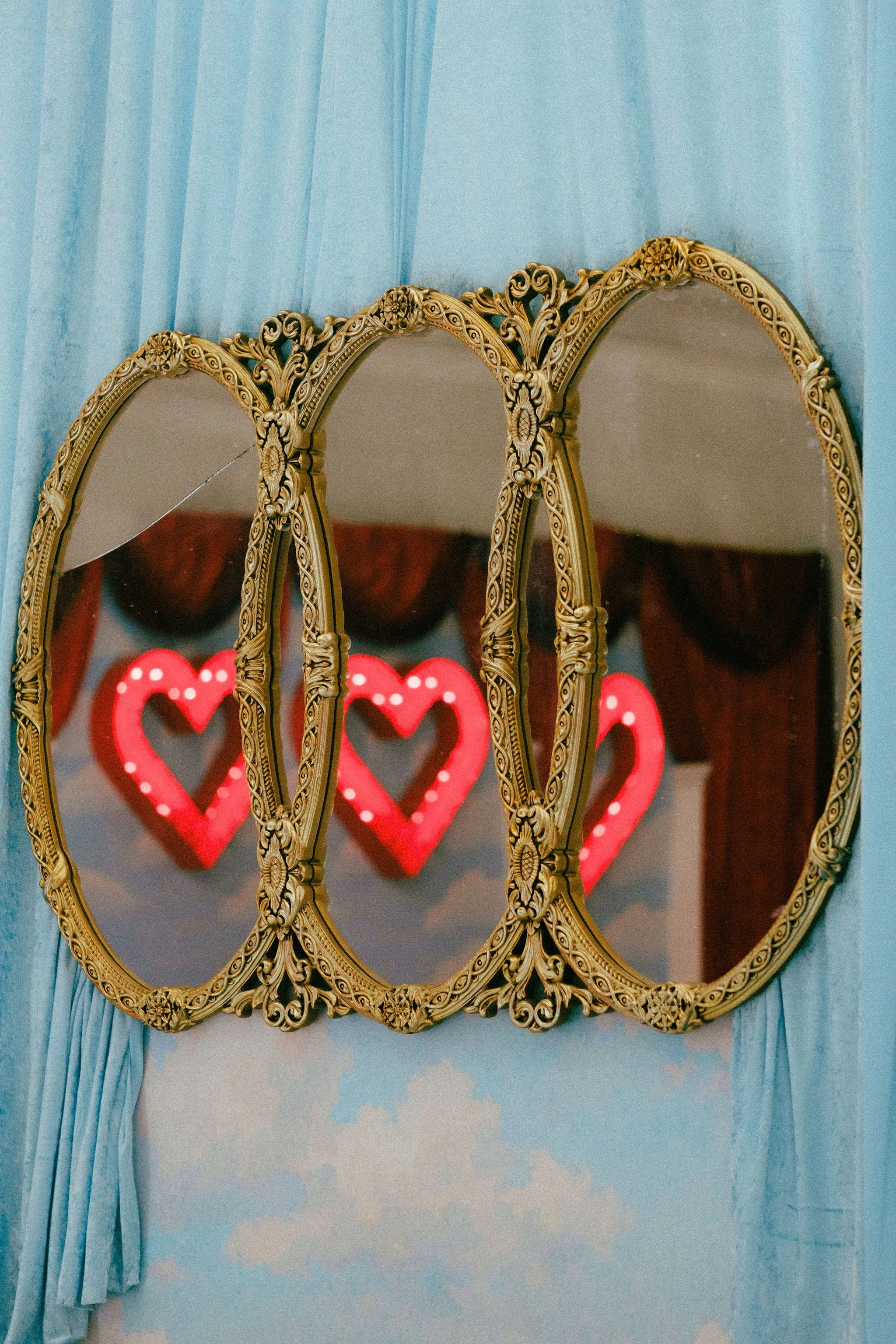 Three ornate gold-framed mirrors hanging on a blue wall, reflecting red neon hearts with a cloudy sky background.