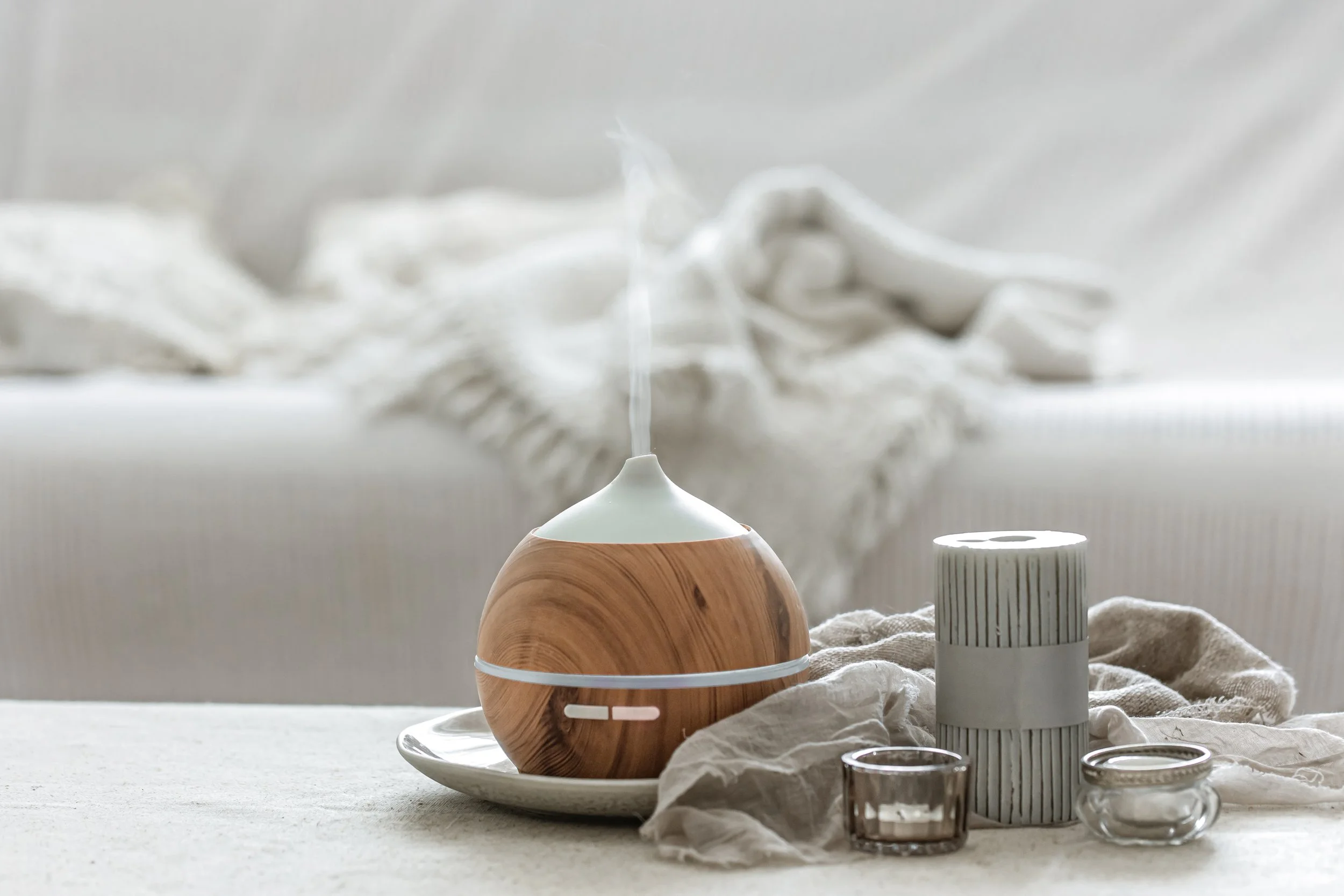 A wooden diffuser on a white plate, surrounded by candles, a glass container, and a fabric cloth, with a neutral background.