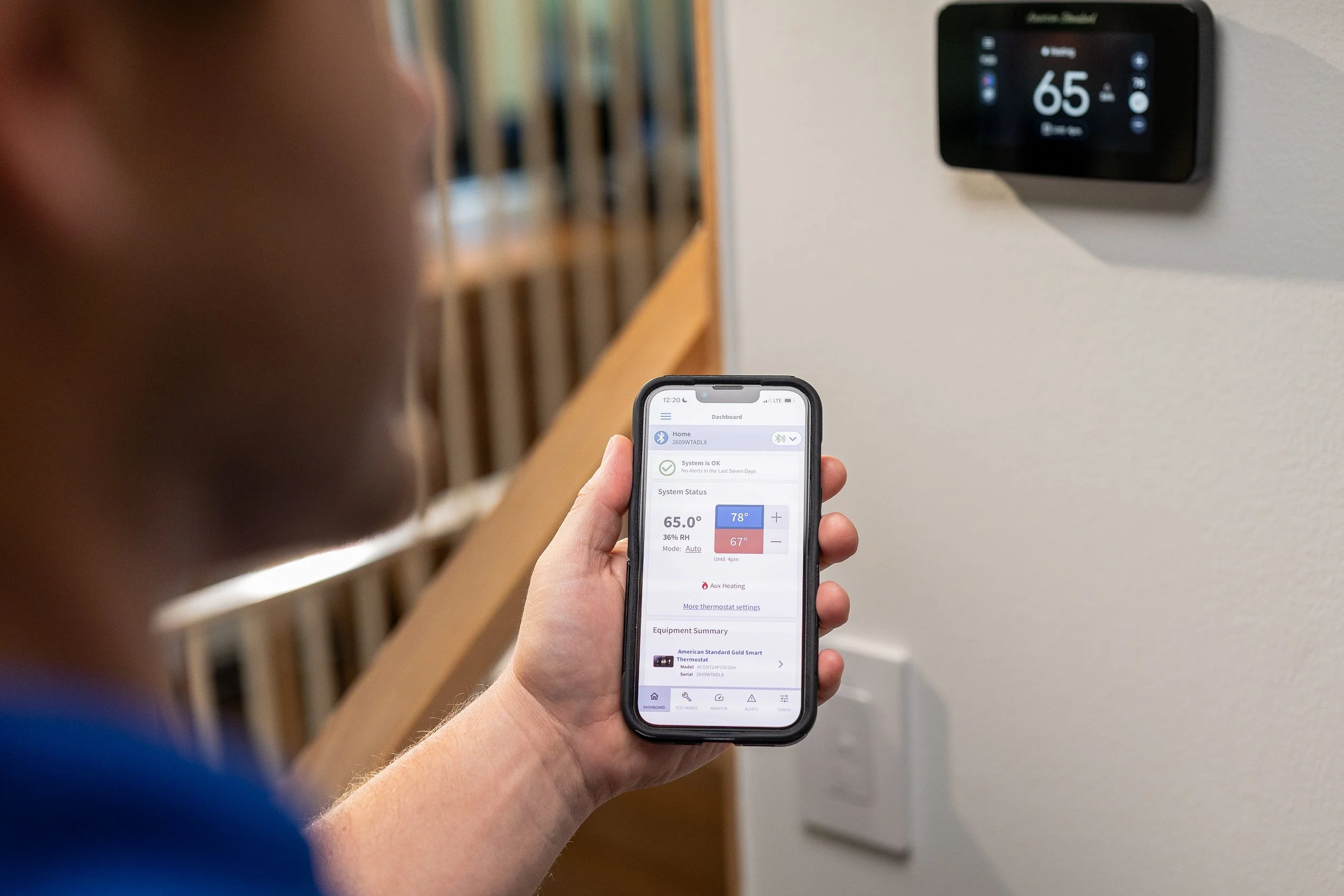 A person holds a smartphone displaying a smart home app, controlling a thermostat on the wall set to 65 degrees Fahrenheit. The background shows part of the person's arm and blurred indoor features.