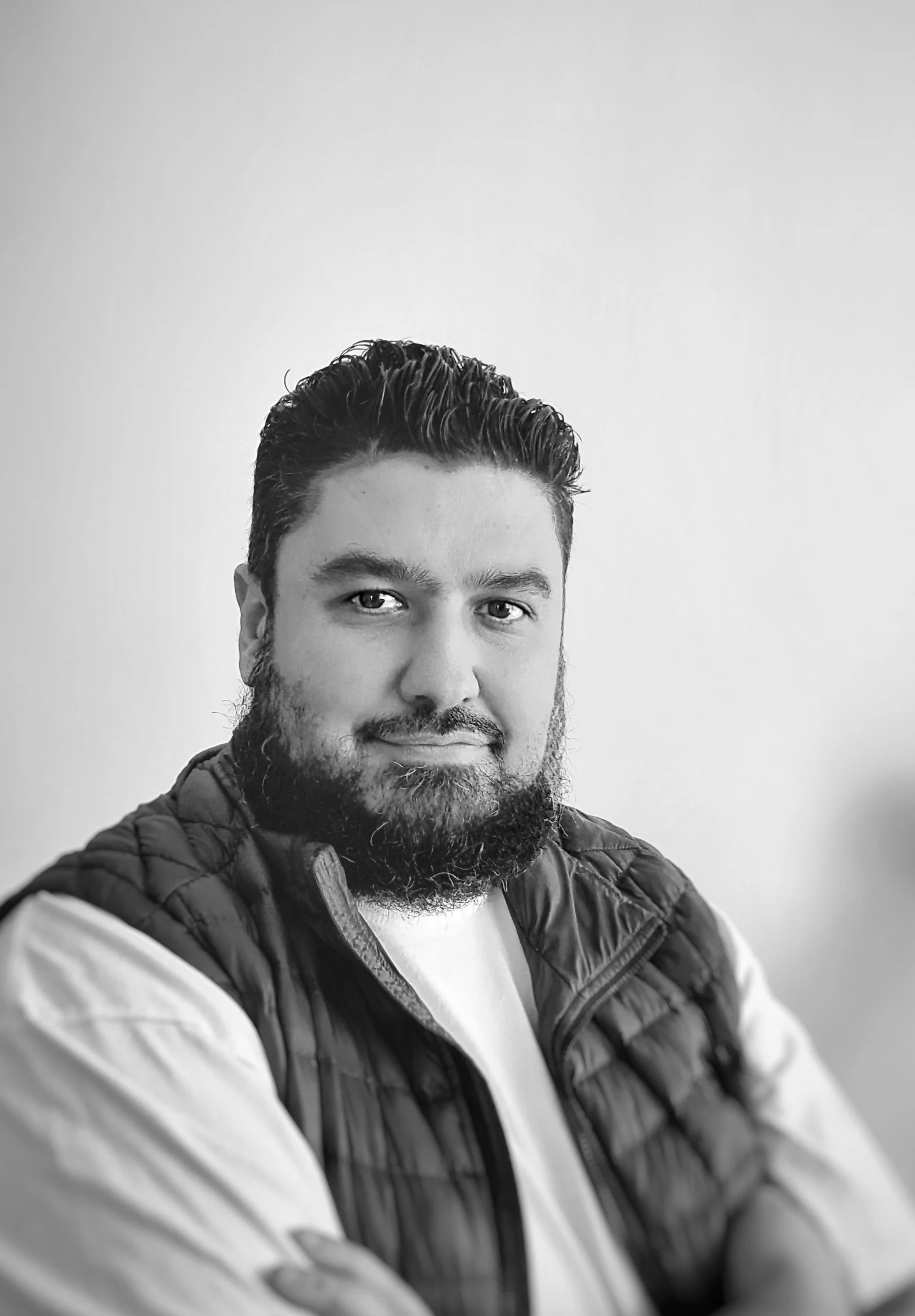 Black and white portrait of a bearded man with styled hair wearing a jacket over a white shirt, smiling slightly.