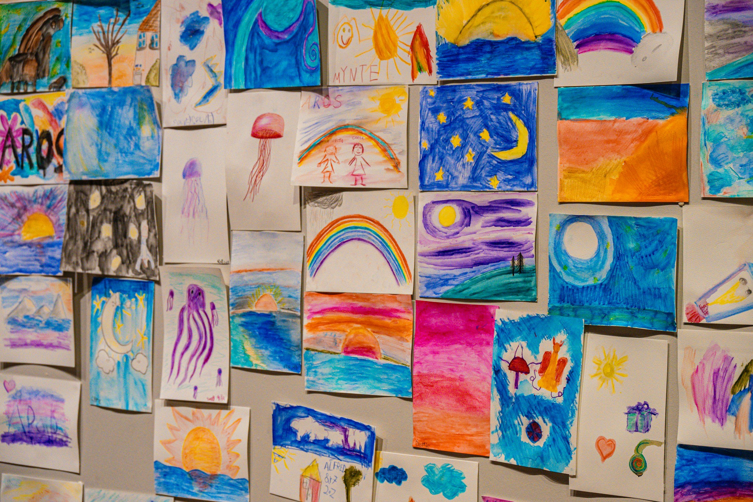 Collection of children's colorful drawings and paintings on a wall, depicting sunsets, rainbows, moons, stars, and various abstract scenes.