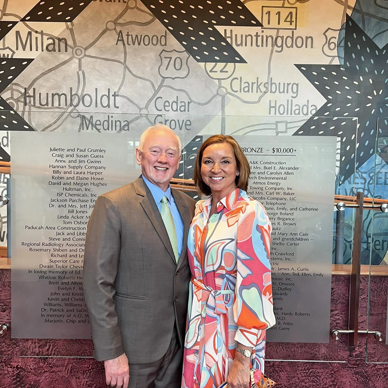 George Bray for McCracken County Judge Executive Kentucky with wife, Angela wearing a colorful, patterned dress at an indoor event. Behind them is a large wall with a map of towns and a list of sponsors or supporters, with black decorative elements.