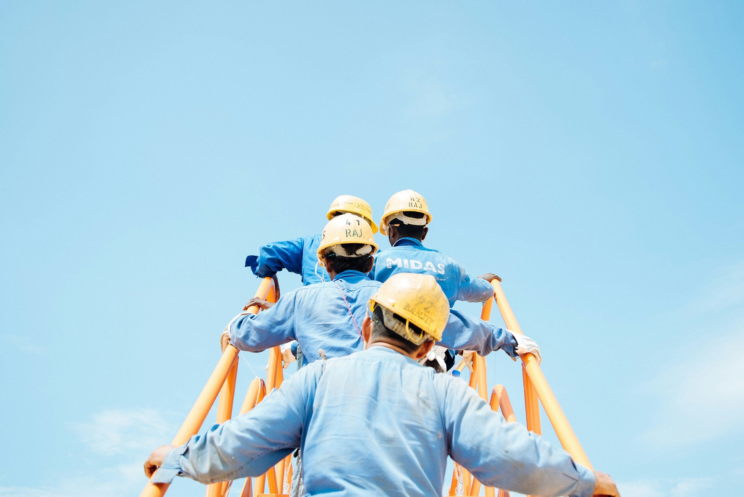 A group of construction workers wearing yellow helmets and blue uniforms climbing a yellow ladder against a clear blue sky.
