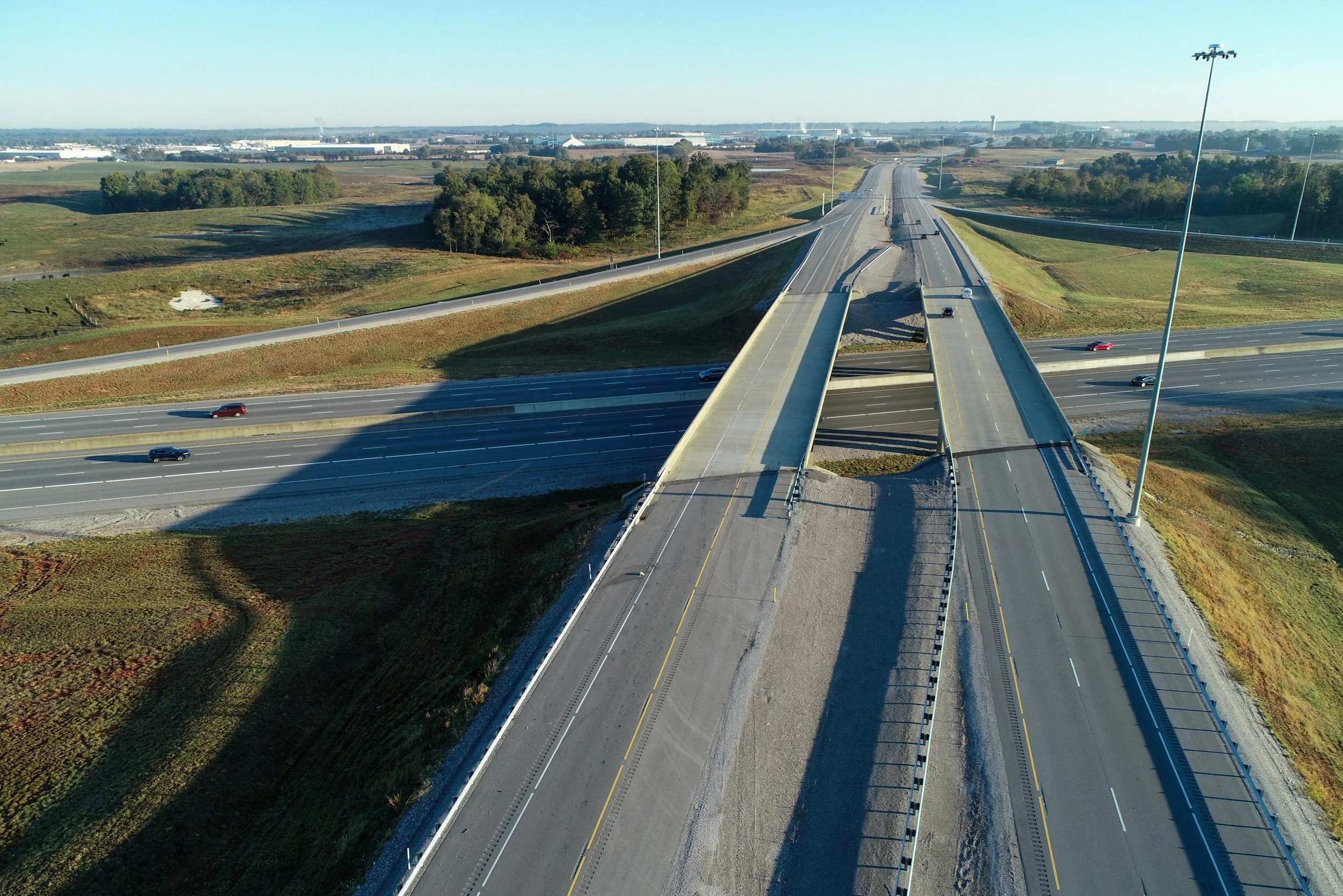 Aerial view of a highway overpass with multiple lanes, cars, and surrounding green landscape under clear blue sky.