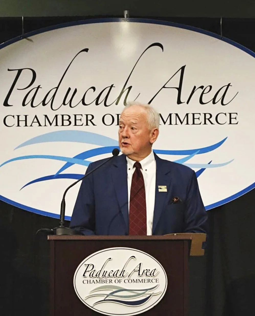 George Bray for McCracken County Judge Executive Kentucky at a podium with a Paducah Area Chamber of Commerce logo. He is speaking into a microphone in front of a large sign that says 'Paducah Area Chamber of Commerce' with stylized blue waves.