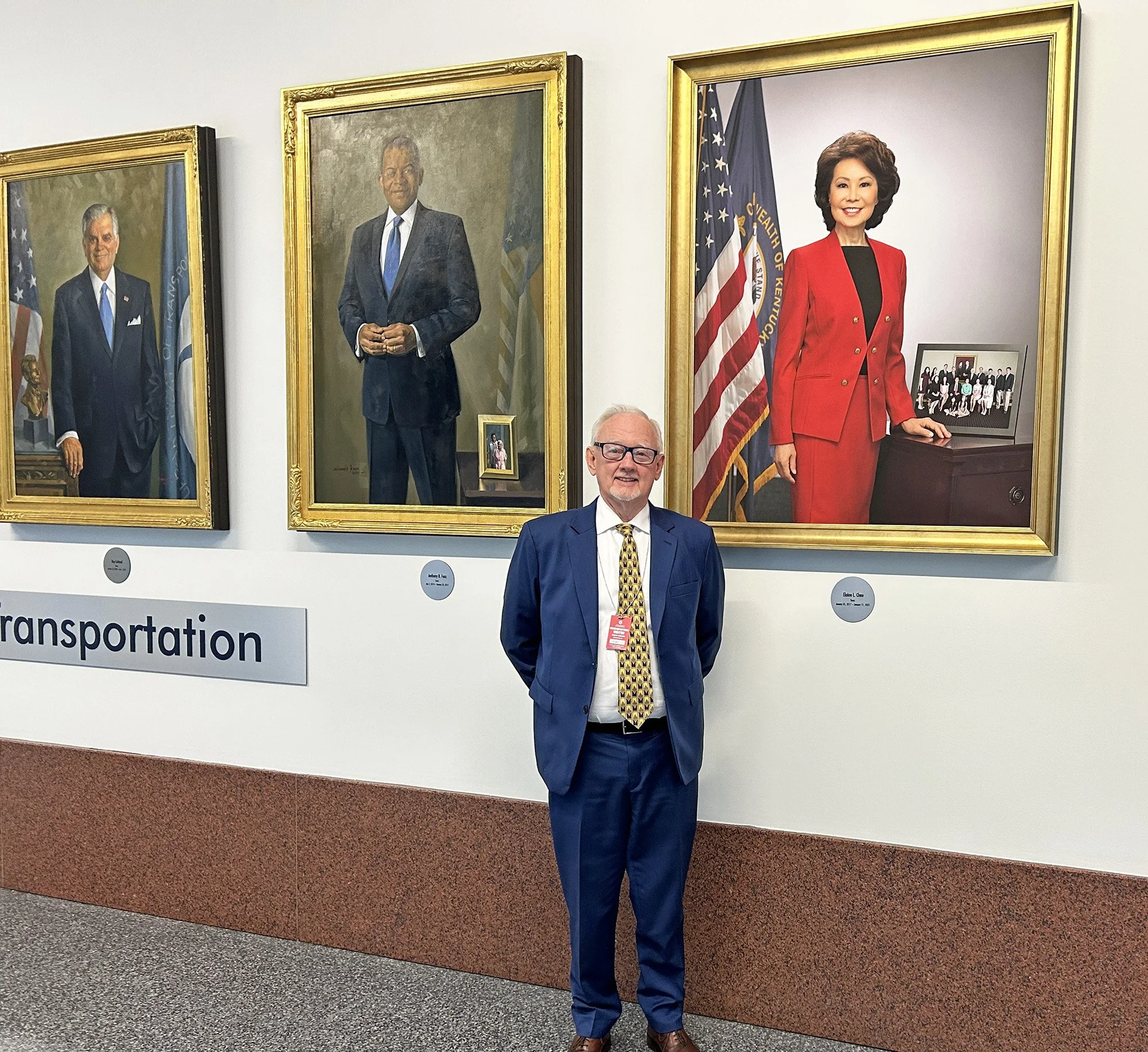 George Bray for McCracken County Judge Executive Kentucky standing in front of framed portraits of political figures, including one of a woman in a red blazer, with American flags in the background.