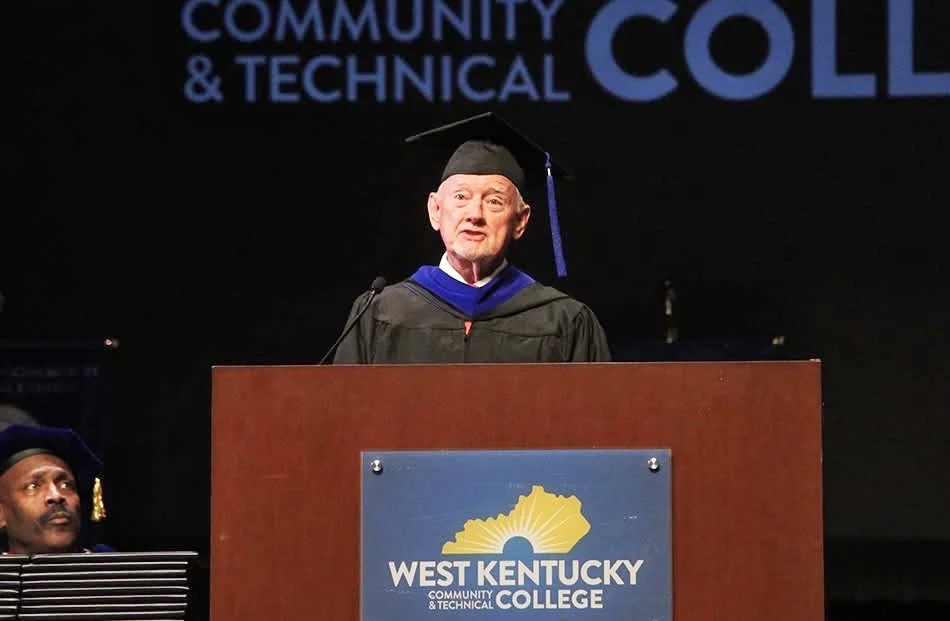 George Bray for McCracken County Judge Executive Kentucky in a graduation cap and gown speaking at a podium during a ceremony at West Kentucky Community & Technical College.