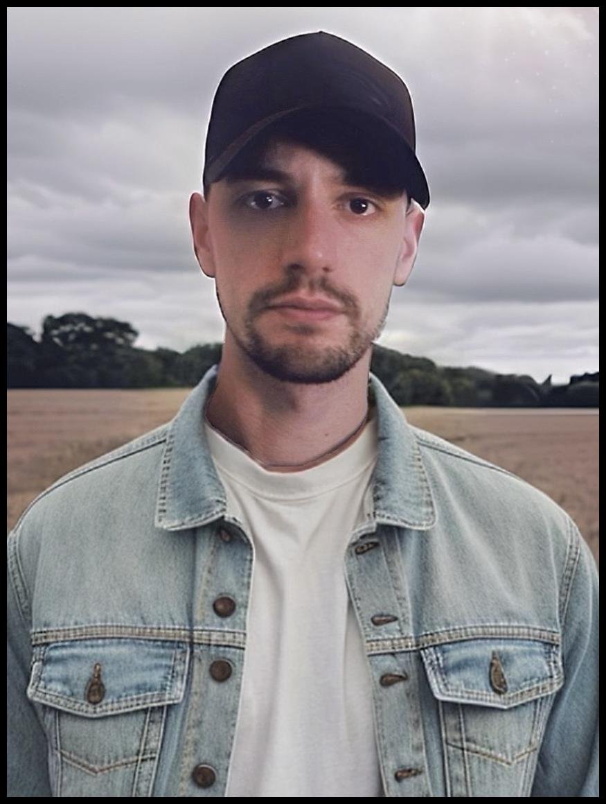 A young man standing outdoors in a field with cloudy skies, wearing a black cap, denim jacket, and white T-shirt.