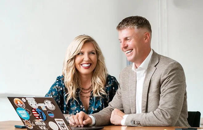Missy Alkema and Rob Alkema smiling and sitting at a table with a laptop covered in stickers, in an office setting.
