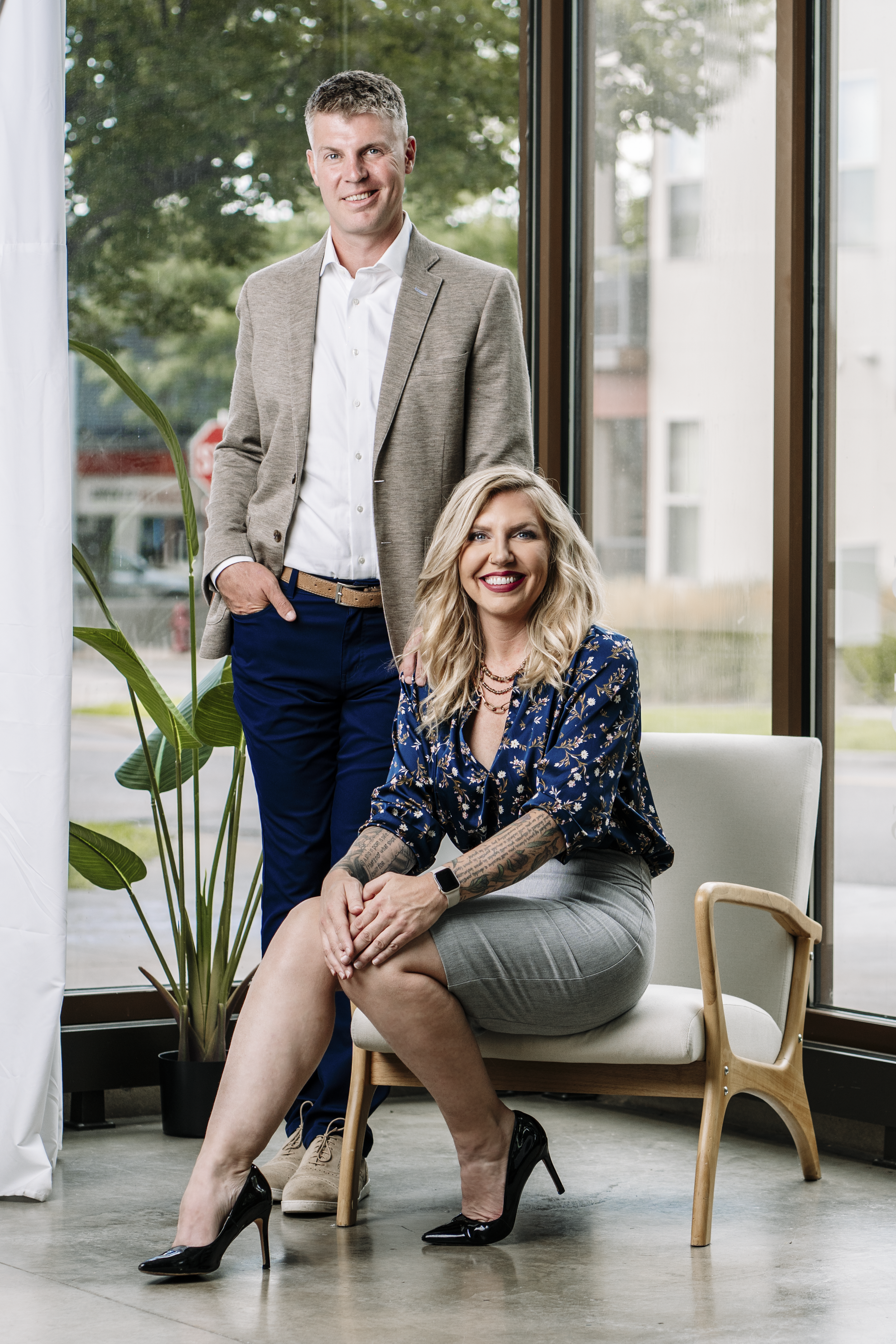Two people in business attire in a modern office setting, one sitting and one standing.