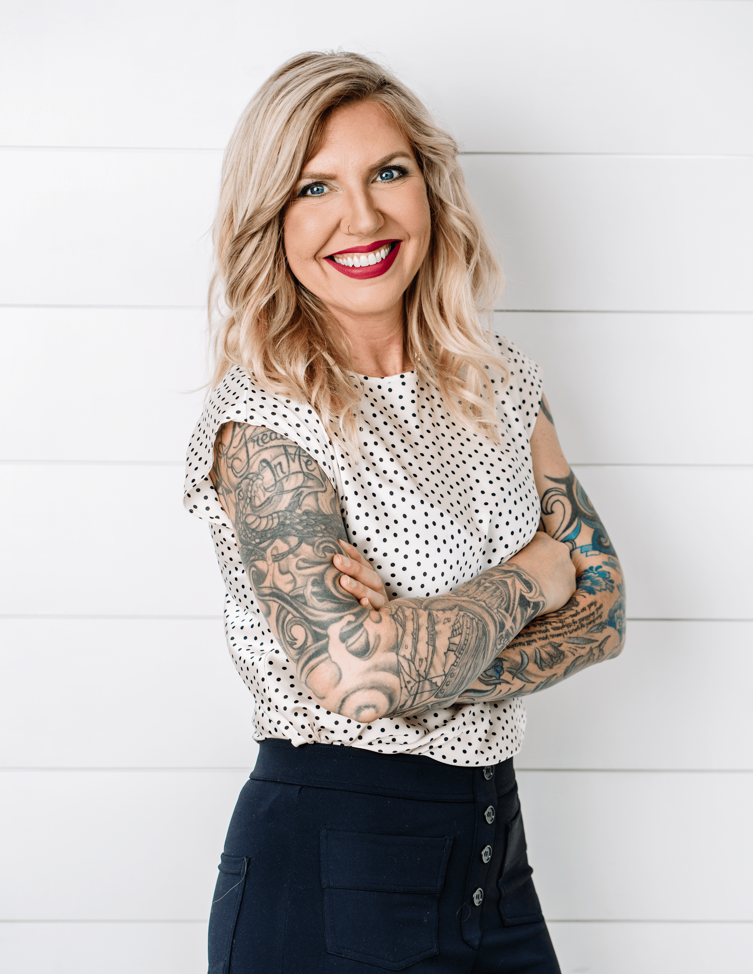 Missy Alkema smiling, wearing polka dot top and dark pants, tattooed arms crossed.