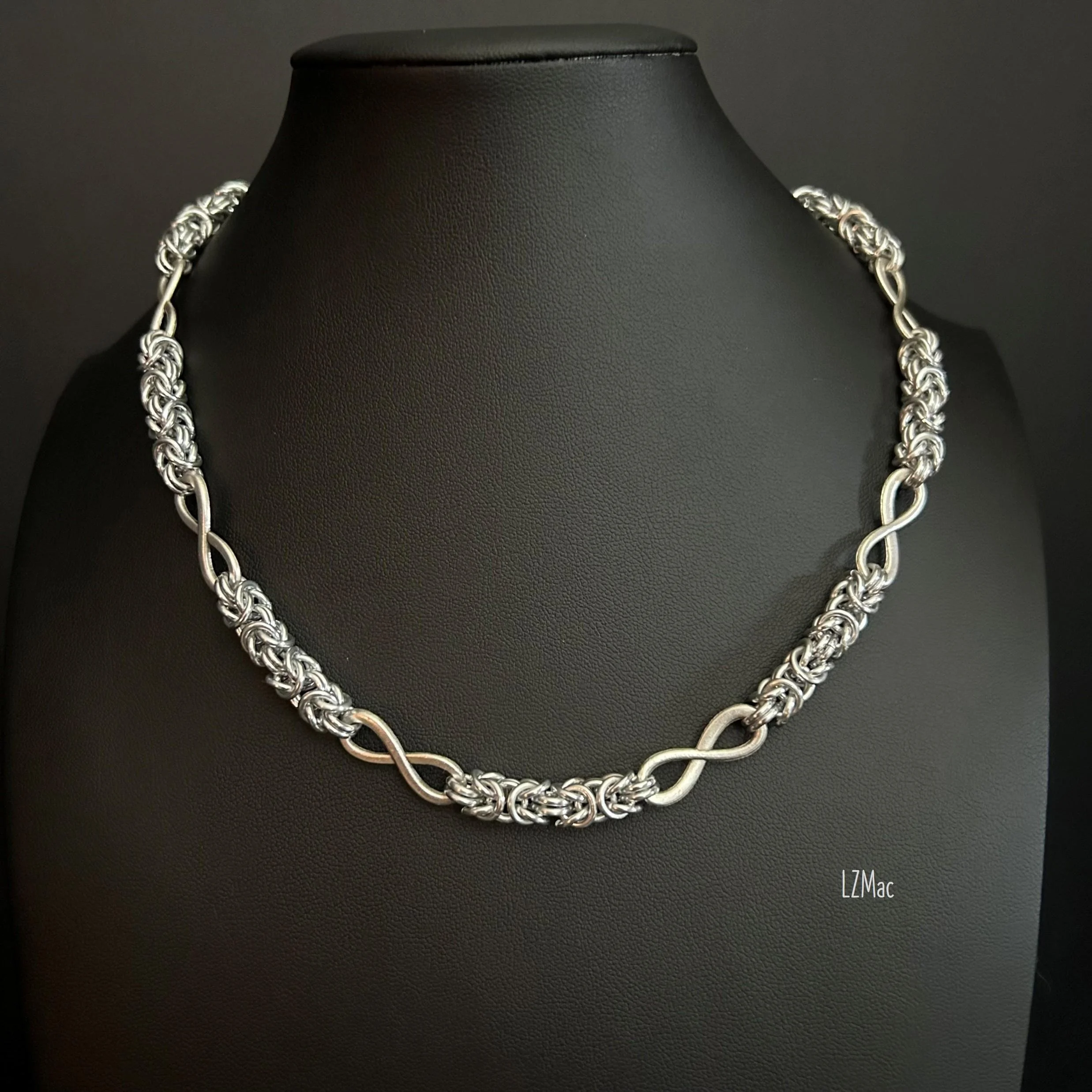 Silver Infinity chainmaille necklace featuring alternating Byzantine weave sections and matte silver infinity connectors displayed on a black jewelry bust.