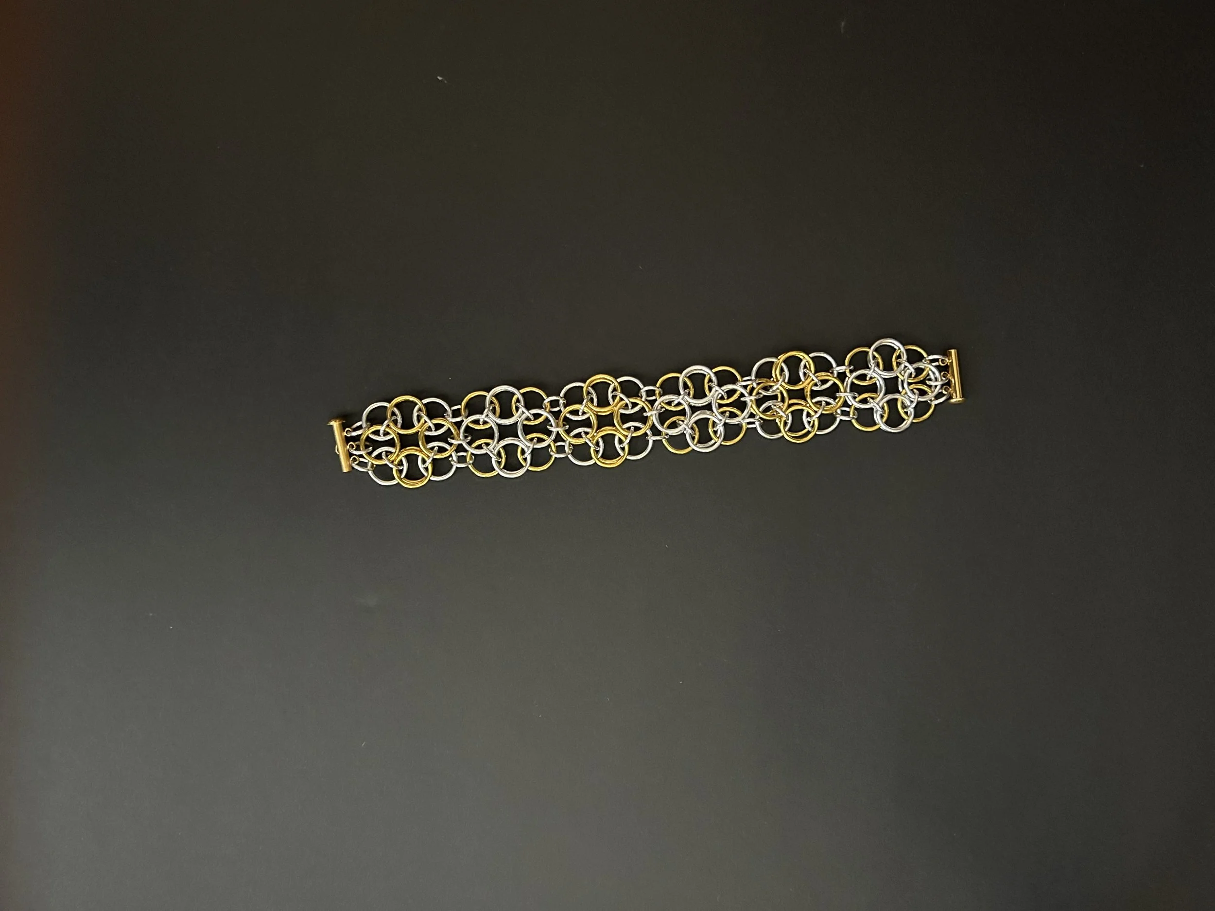 Chainmaille bracelet in sliver- and gold-tone helm rosette weave  laid flat on black background