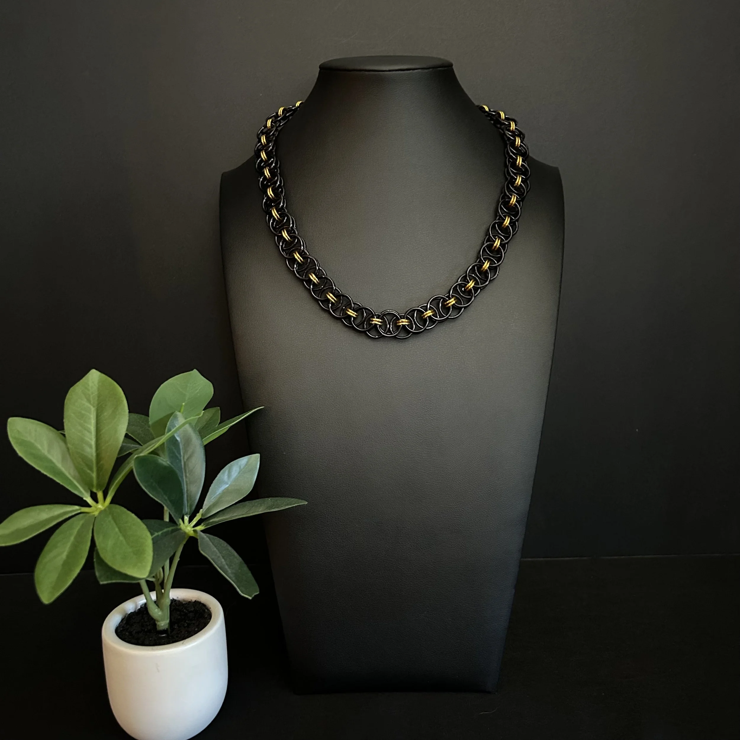 Blackgold Ladder necklace shown in full length beside a green plant, highlighting its black and gold aluminum rings and modern chainmaille design.