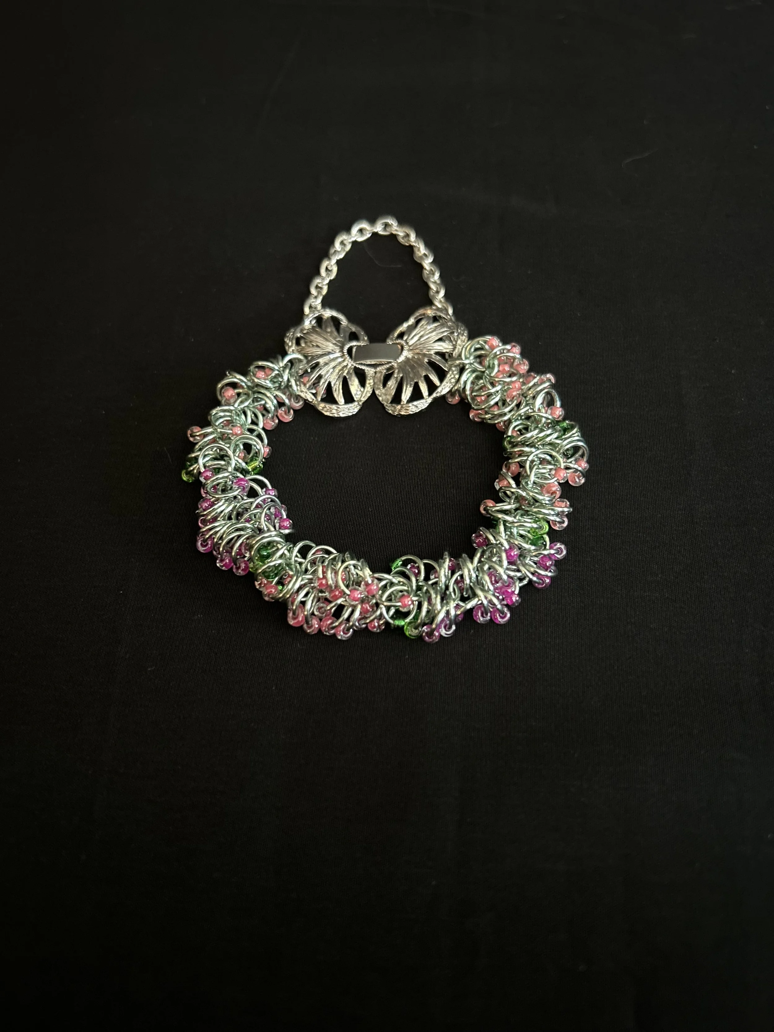 Chainmaille bracelet arranged in a circle on a black background, showing the butterfly clasp, safety chain, and pink and green beadwork.