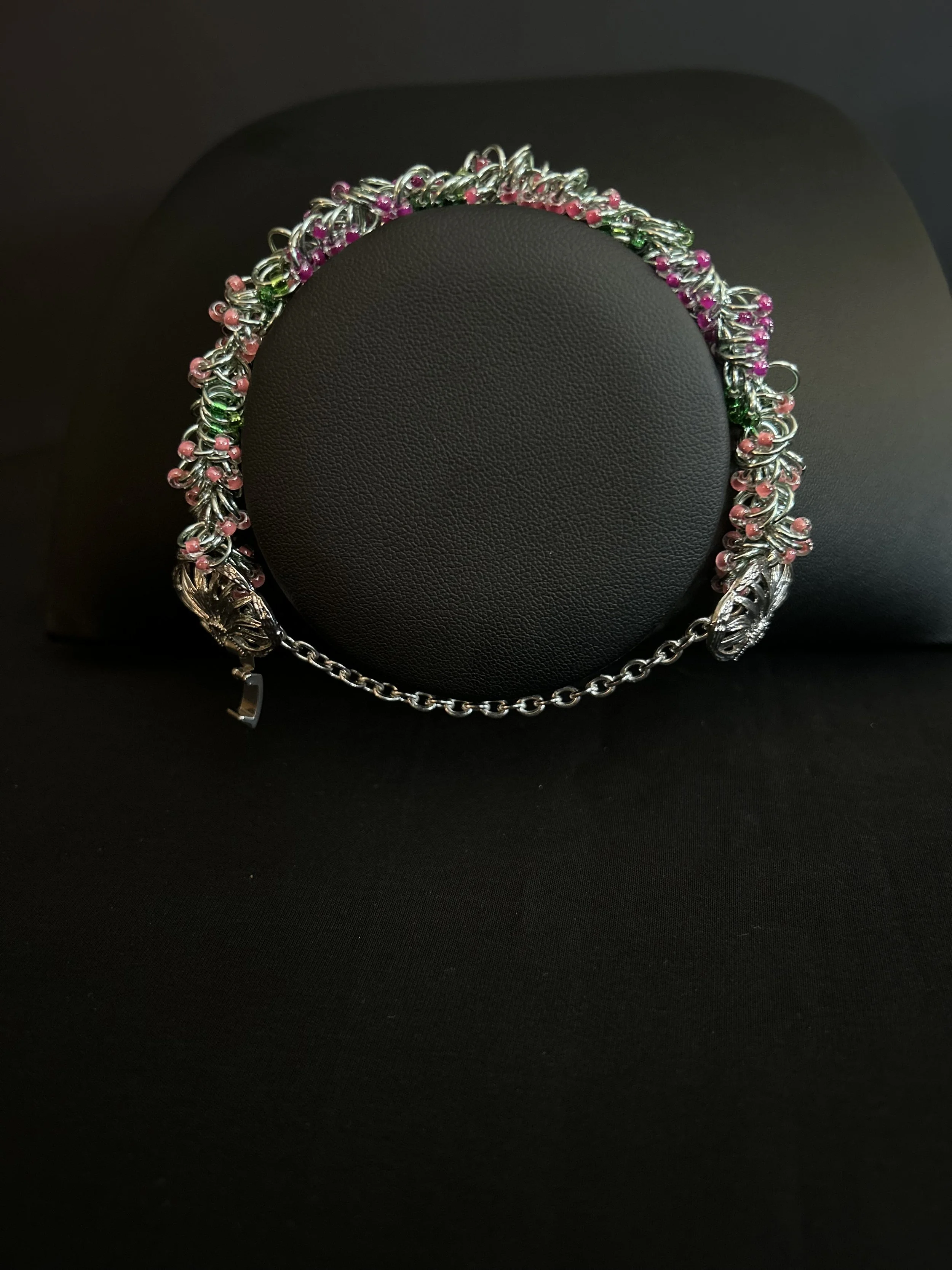Chainmaille bracelet on a black cylindrical display, highlighting the single‑strand weave, pink and green 8/0 Miyuki beads, and green‑tinged silver rings.