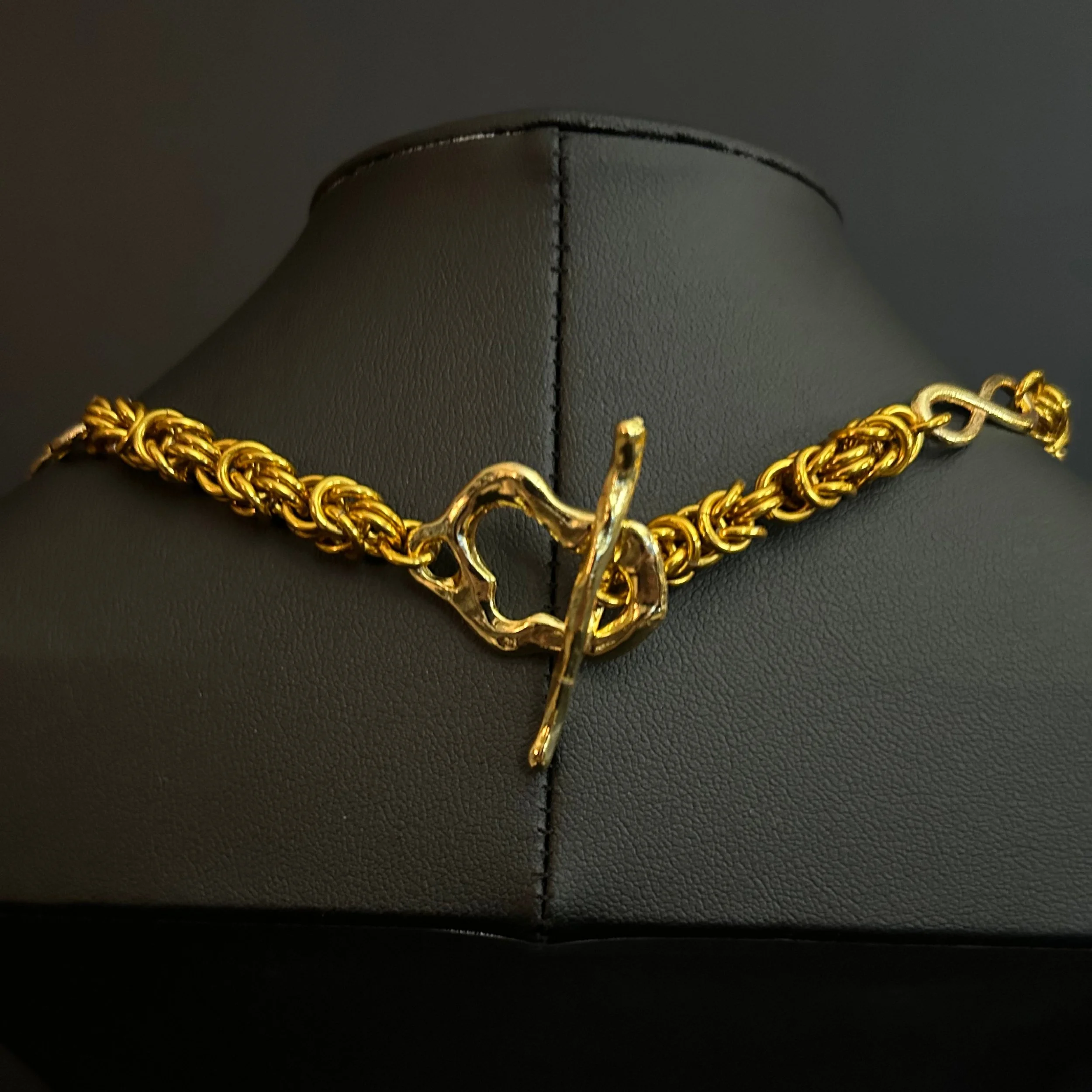 Close-up of the Golden Infinity necklace showing the bright gold Byzantine weave and sculptural toggle clasp on a black display