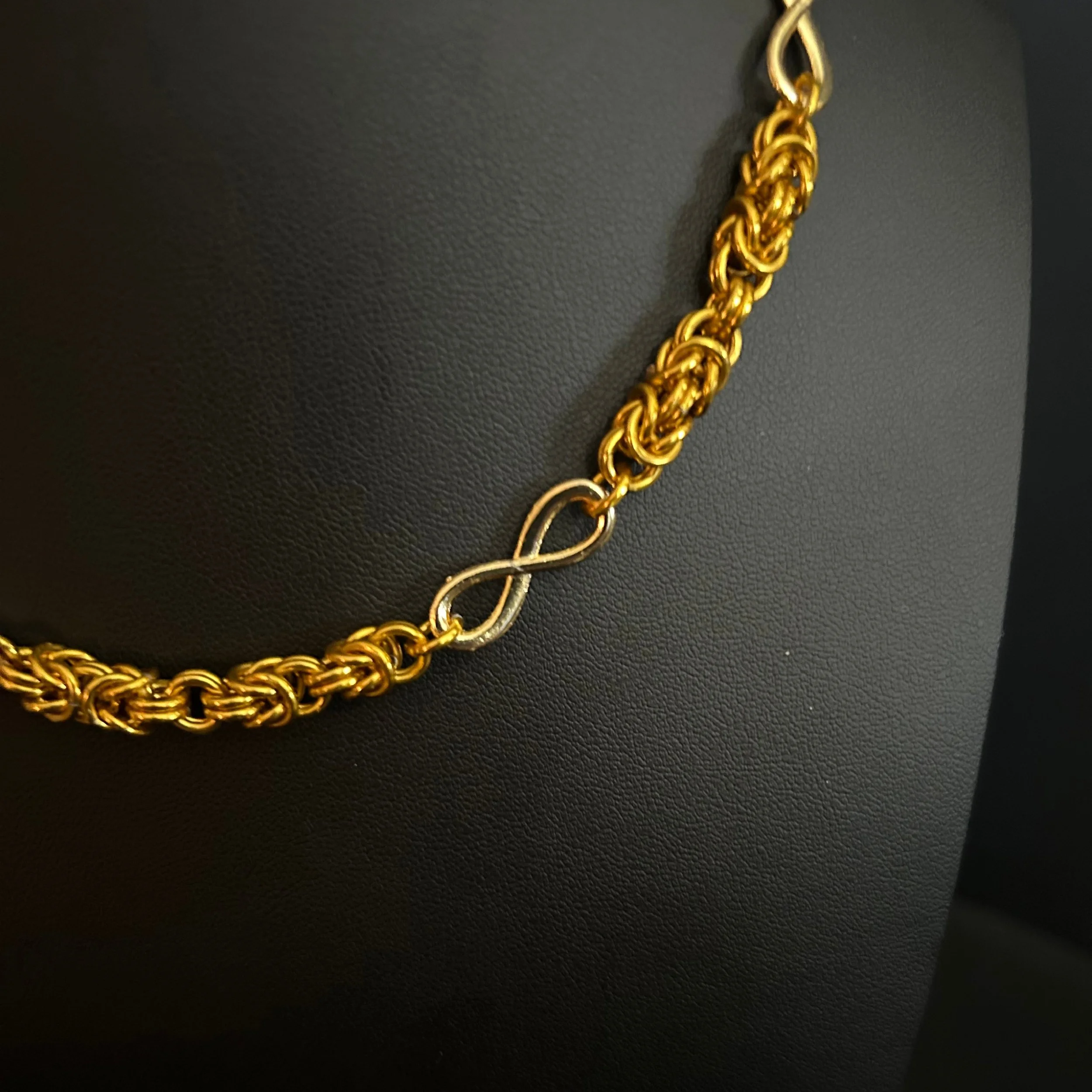 Golden Infinity necklace with a central gold infinity connector and polished Byzantine links, shown on a dark jewelry stand