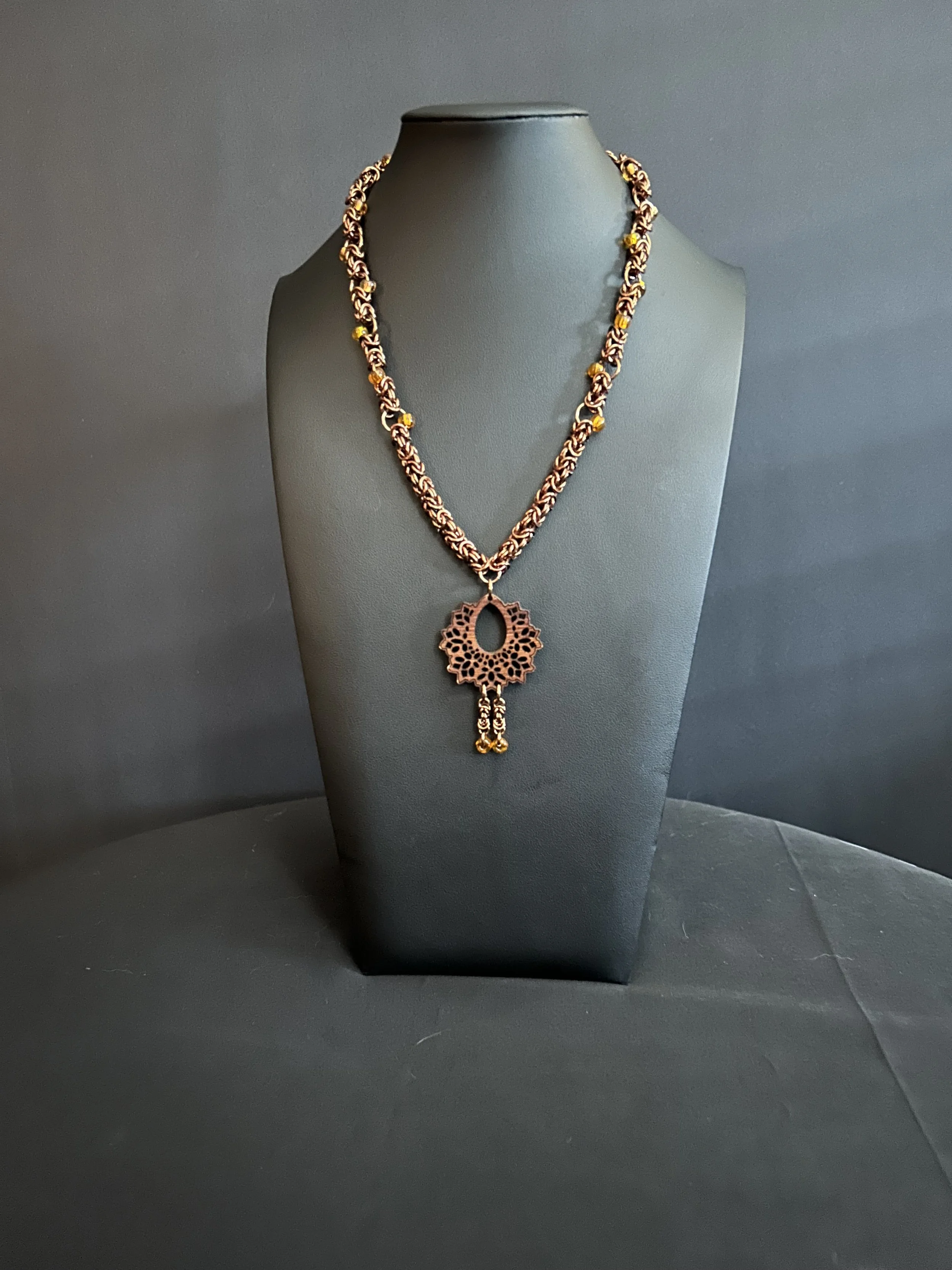 Full view of the necklace on a black bust, showing the bronze chain with amber beads, the ornate wood pendant, and the small dangling bead accents.