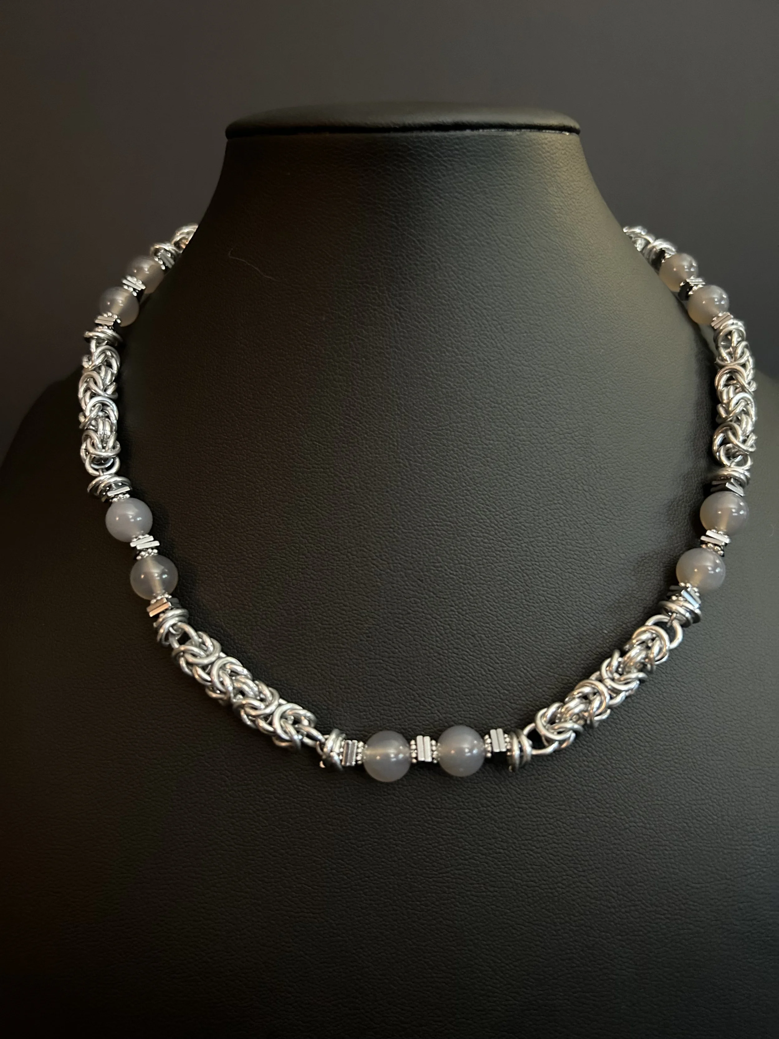 Moonlit Harbor necklace displayed on a black jewelry bust, featuring gray chalcedony beads, hematite spacers, and handwoven aluminum chainmaille.