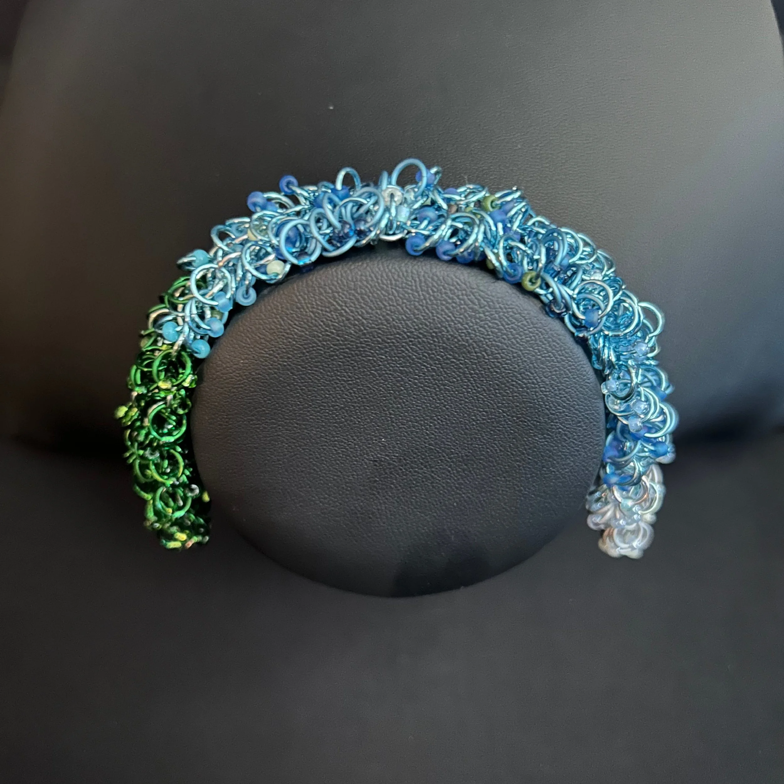 “Mountain‑tone chainmaille bracelet with green and blue beads displayed on a black jewelry bust.”