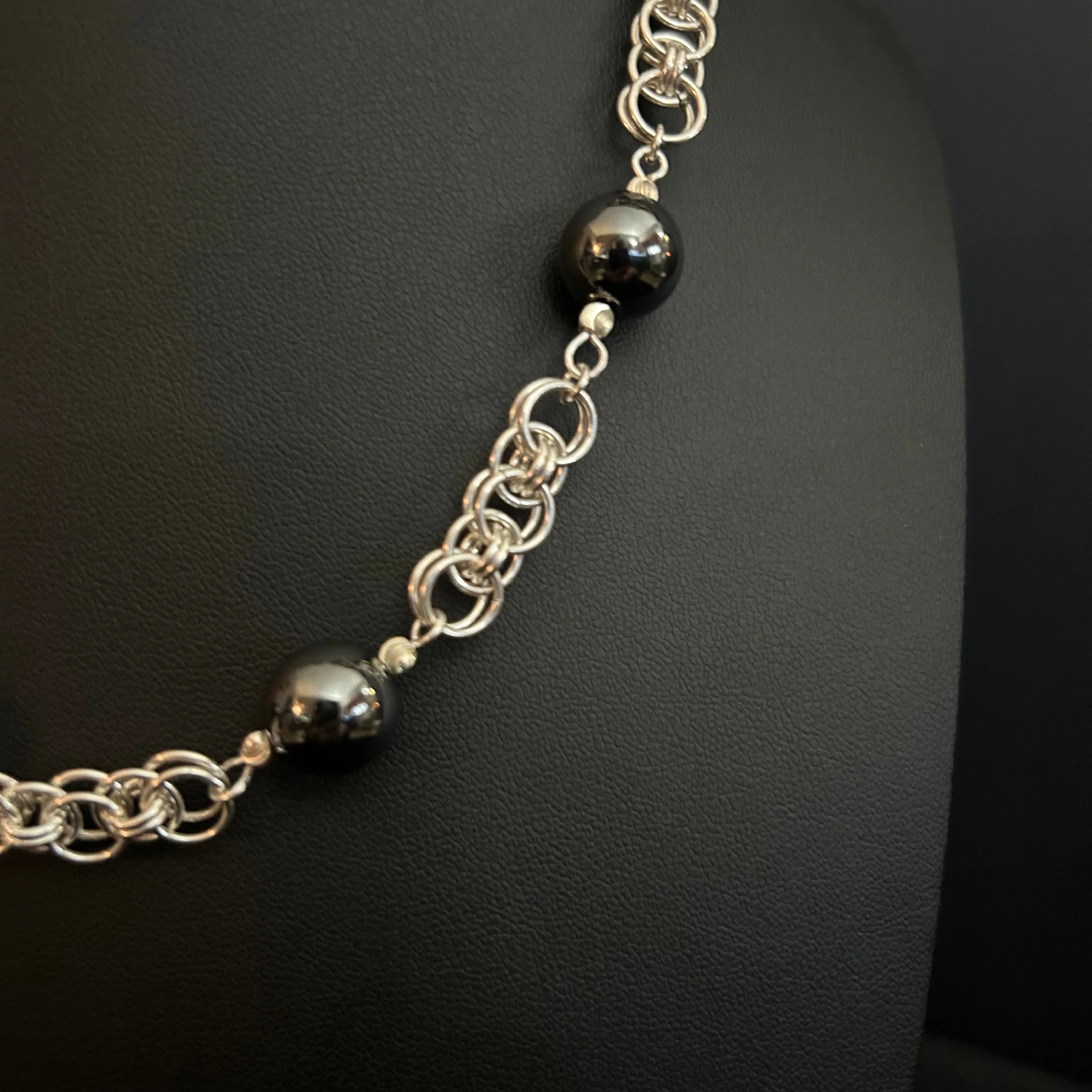 Detail of the Silver Orbit necklace with sterling helm chainmaille links and glossy hematite beads against a black background.
