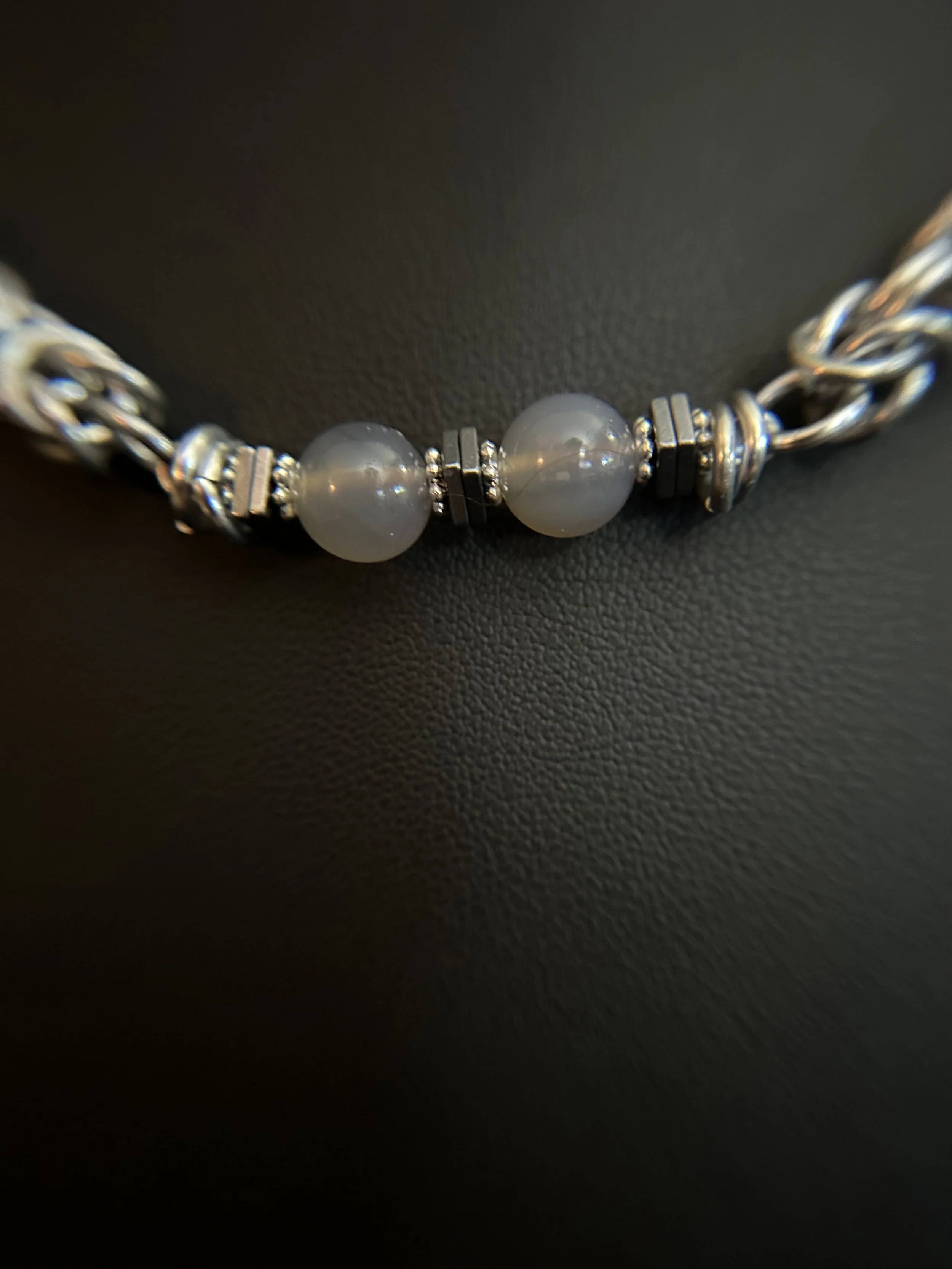 Close-up of a gray chalcedony bead framed by square hematite spacers, connected to bright handwoven aluminum chainmaille.