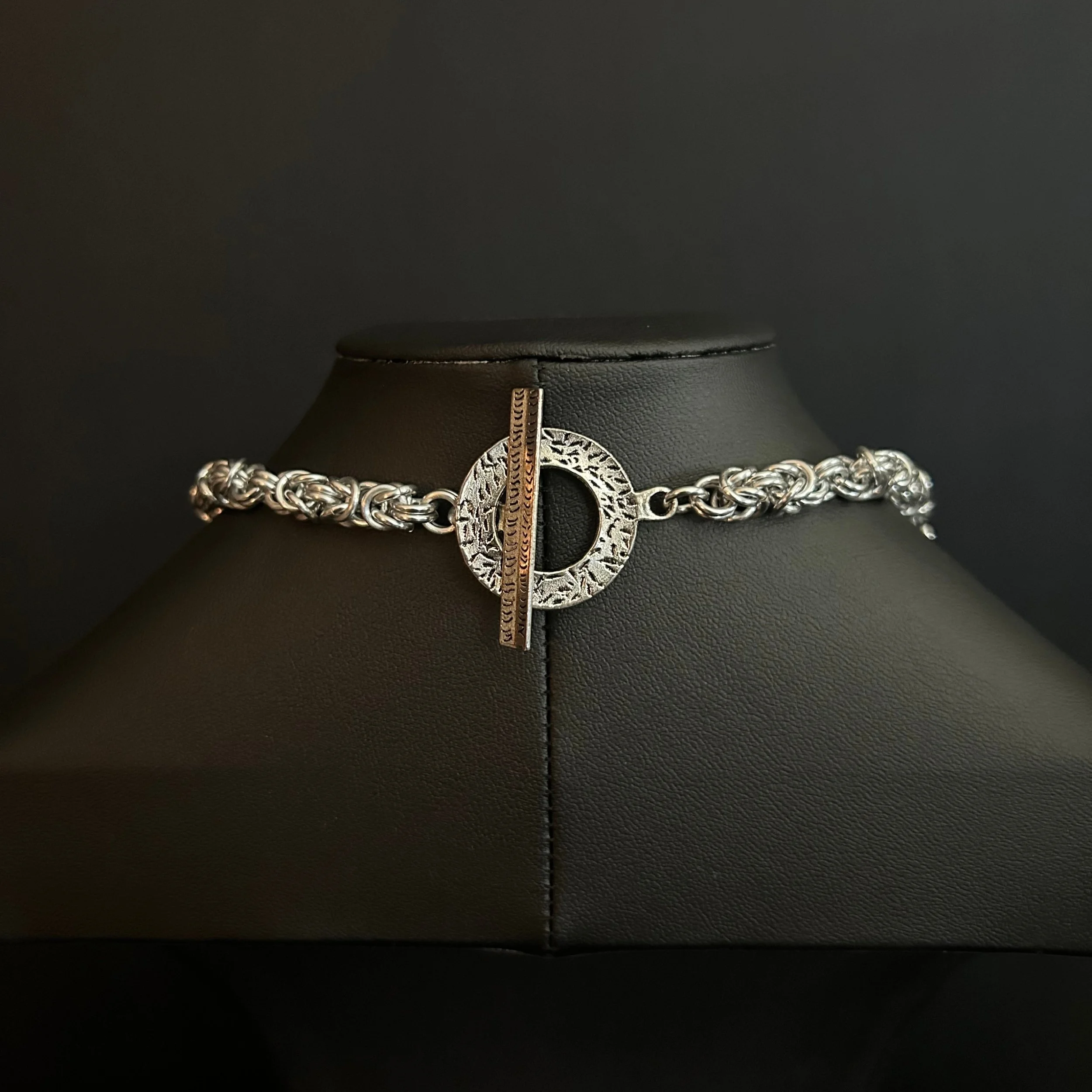 Silver Forest bracelet with interwoven silver links and a textured toggle clasp, displayed on a black stand