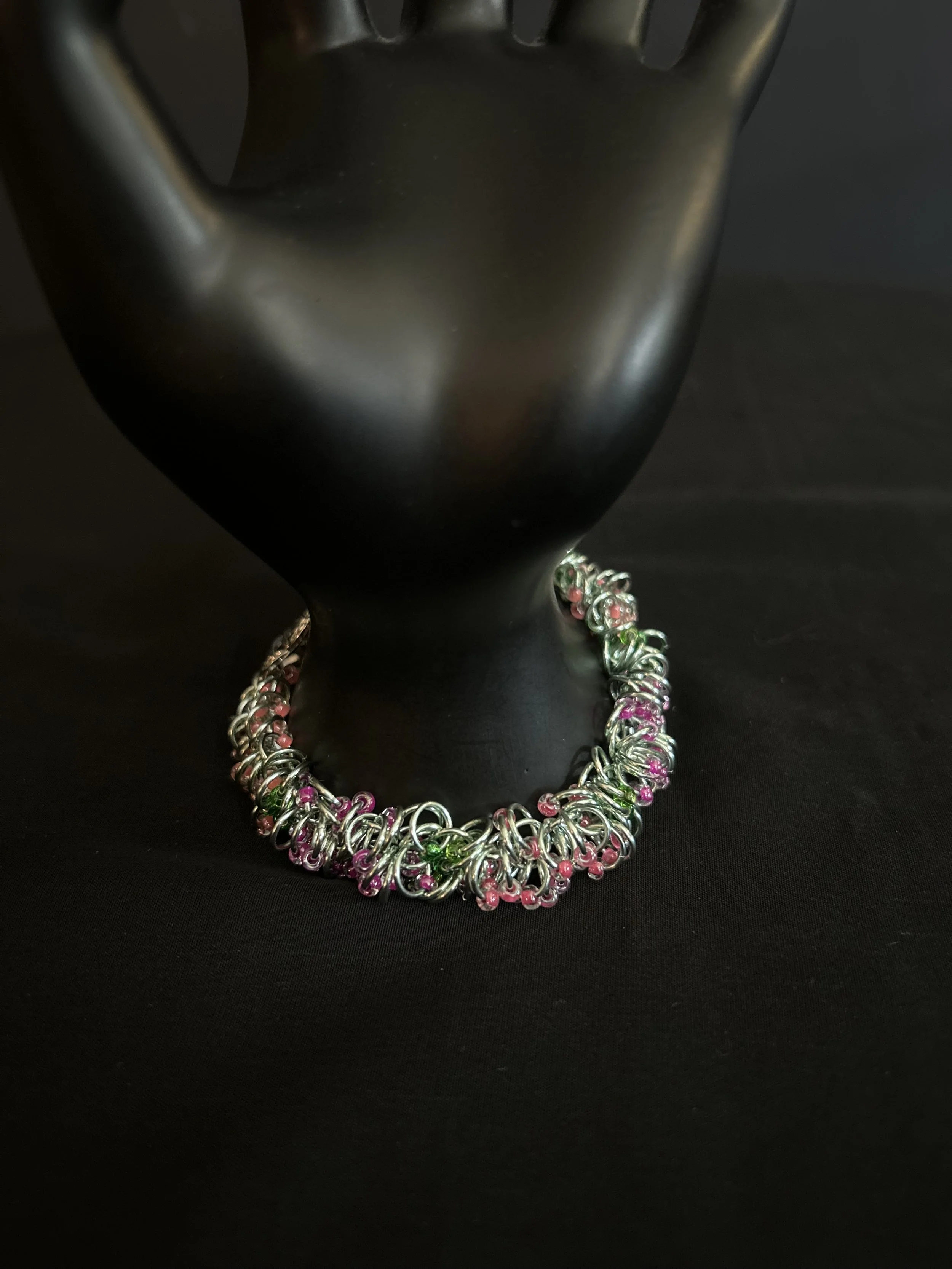 Chainmaille bracelet displayed on a black mannequin hand, showing the single‑strand weave, pink and green beads, and silver‑plated clasp.