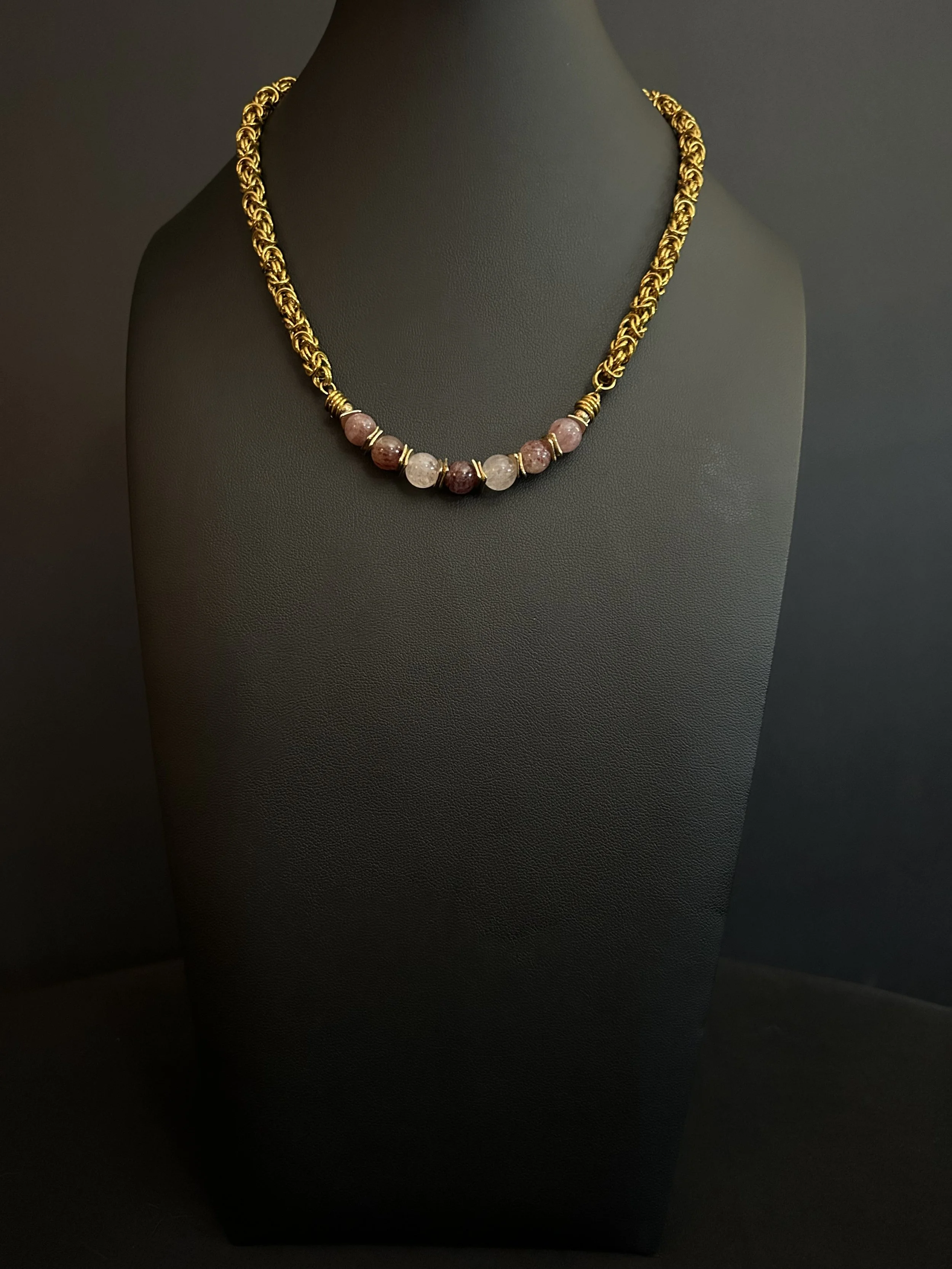 Hand‑woven gold‑tone chainmaille necklace with seven 8mm strawberry quartz beads and gold‑plated spacers, displayed on a black jewelry stand.