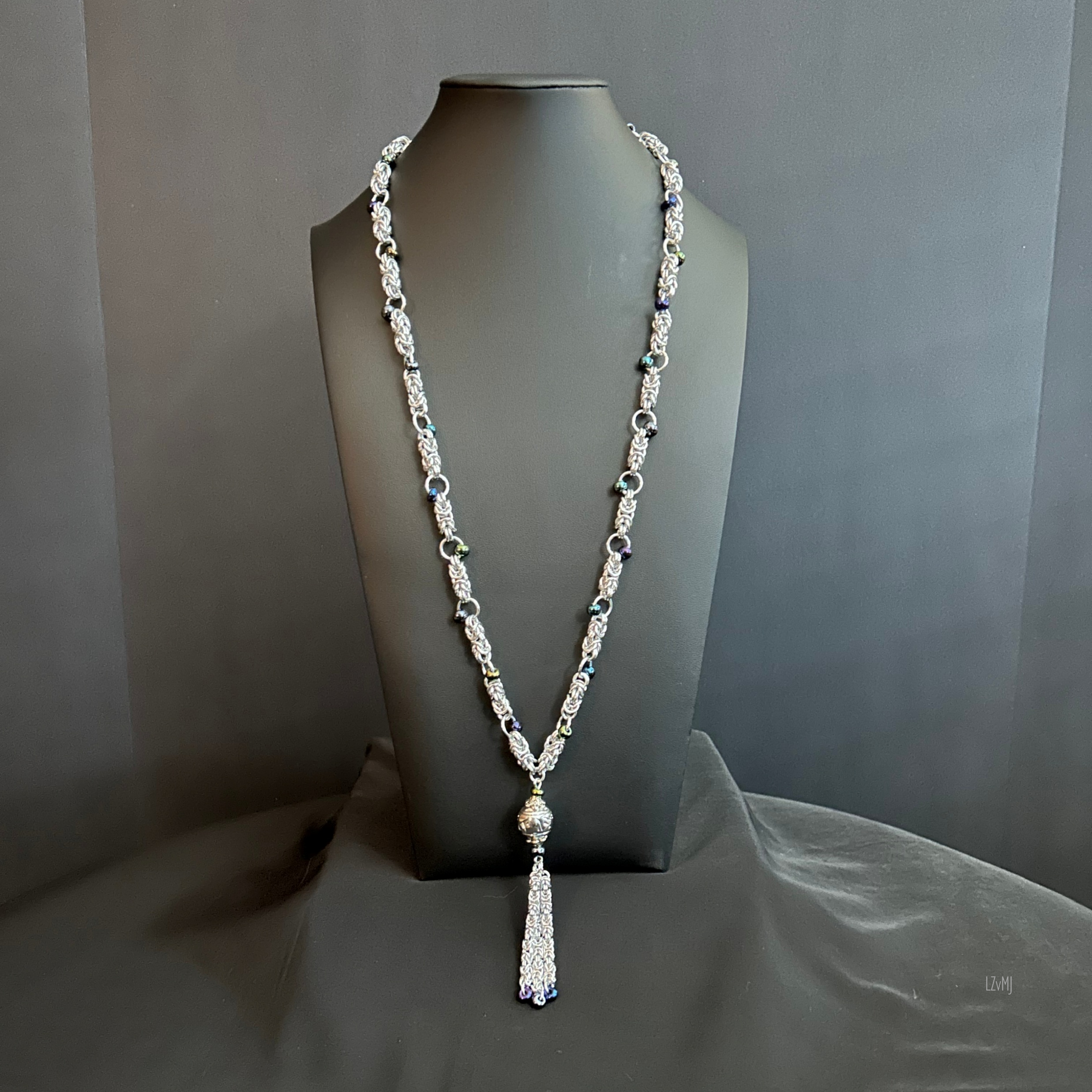 Midnight Tassel necklace shown in full length on a black bust, featuring silver chainmaille, dark beads, a patterned central bead, and a long chain tassel.