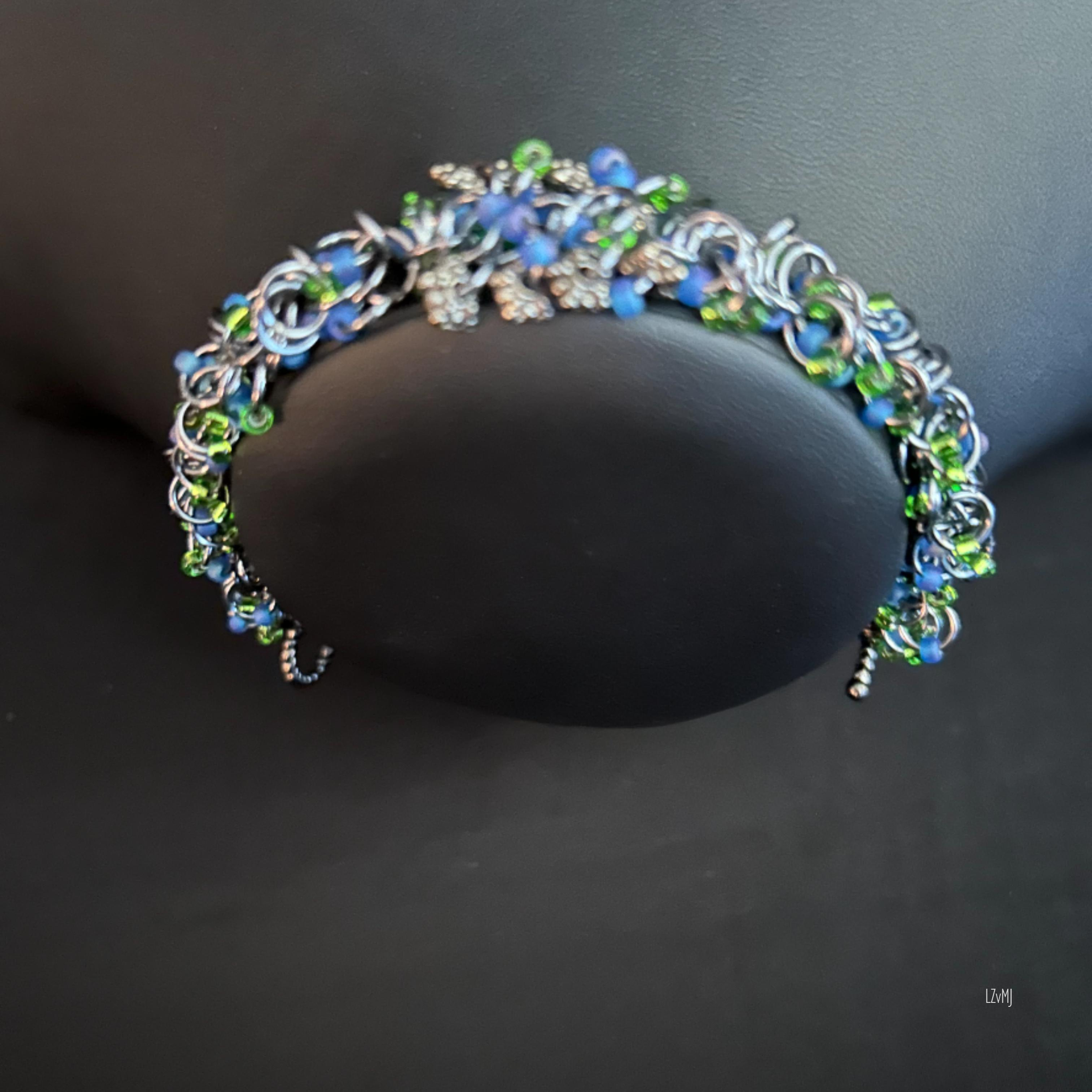 “Black Ice chainmaille bracelet with blue and green beads displayed on a black bust.”