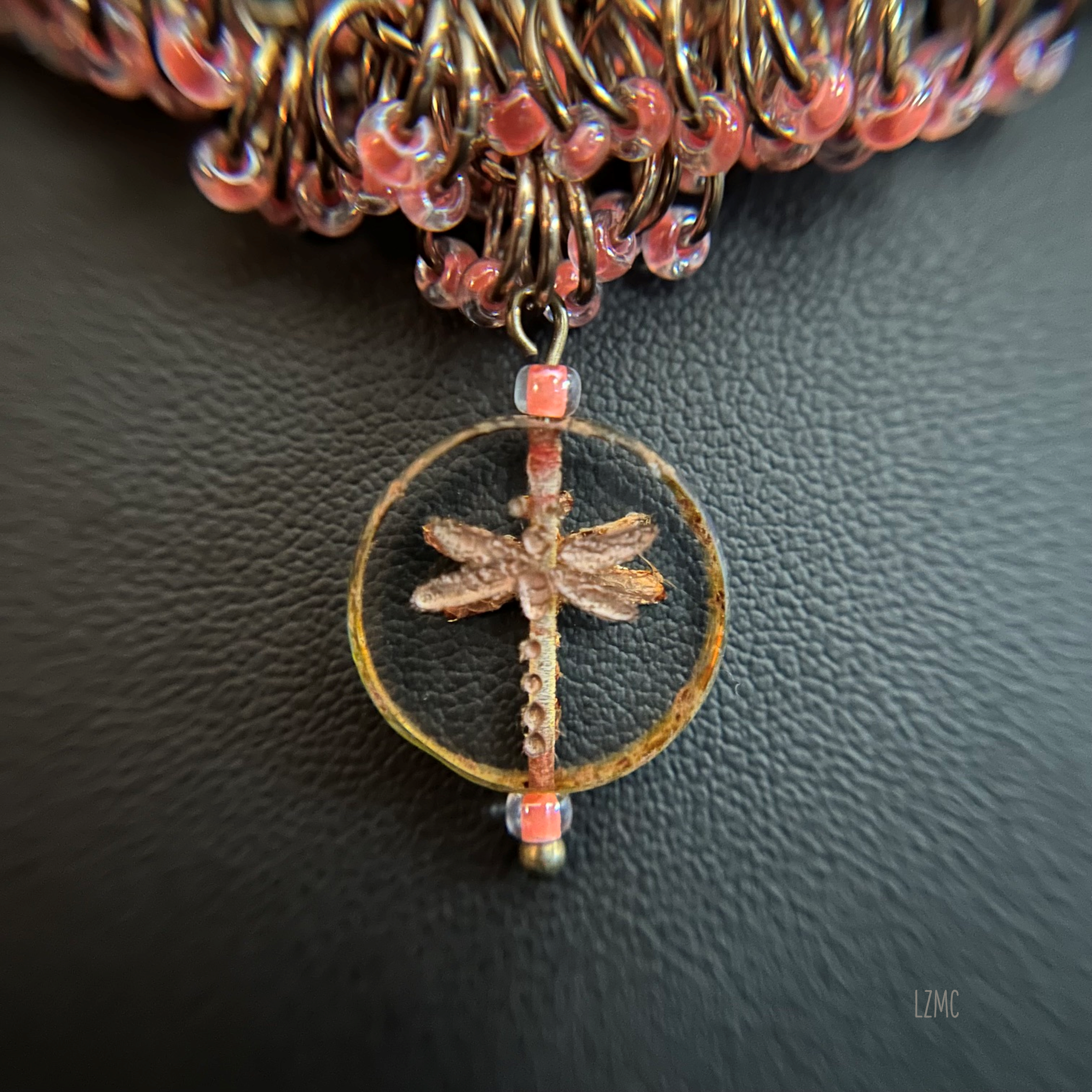 Close‑up of the Czech Glass Bronzed Crystal Picasso dragonfly coin focal, highlighting its bronze shimmer and detailed glass texture within the champagne and pink chainmaille weave.