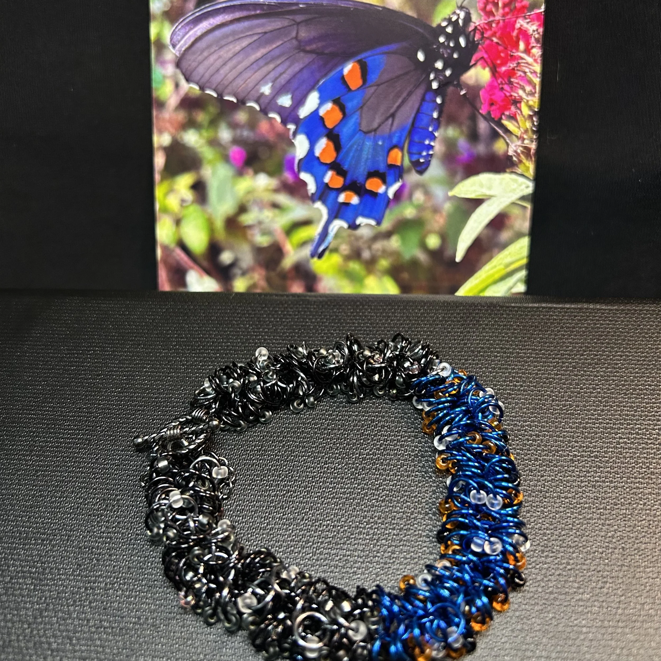 A flat‑laid shaggy‑loop bracelet in black, Black Ice, and royal blue aluminum rings, shown beside a butterfly photo that inspired the color palette.
