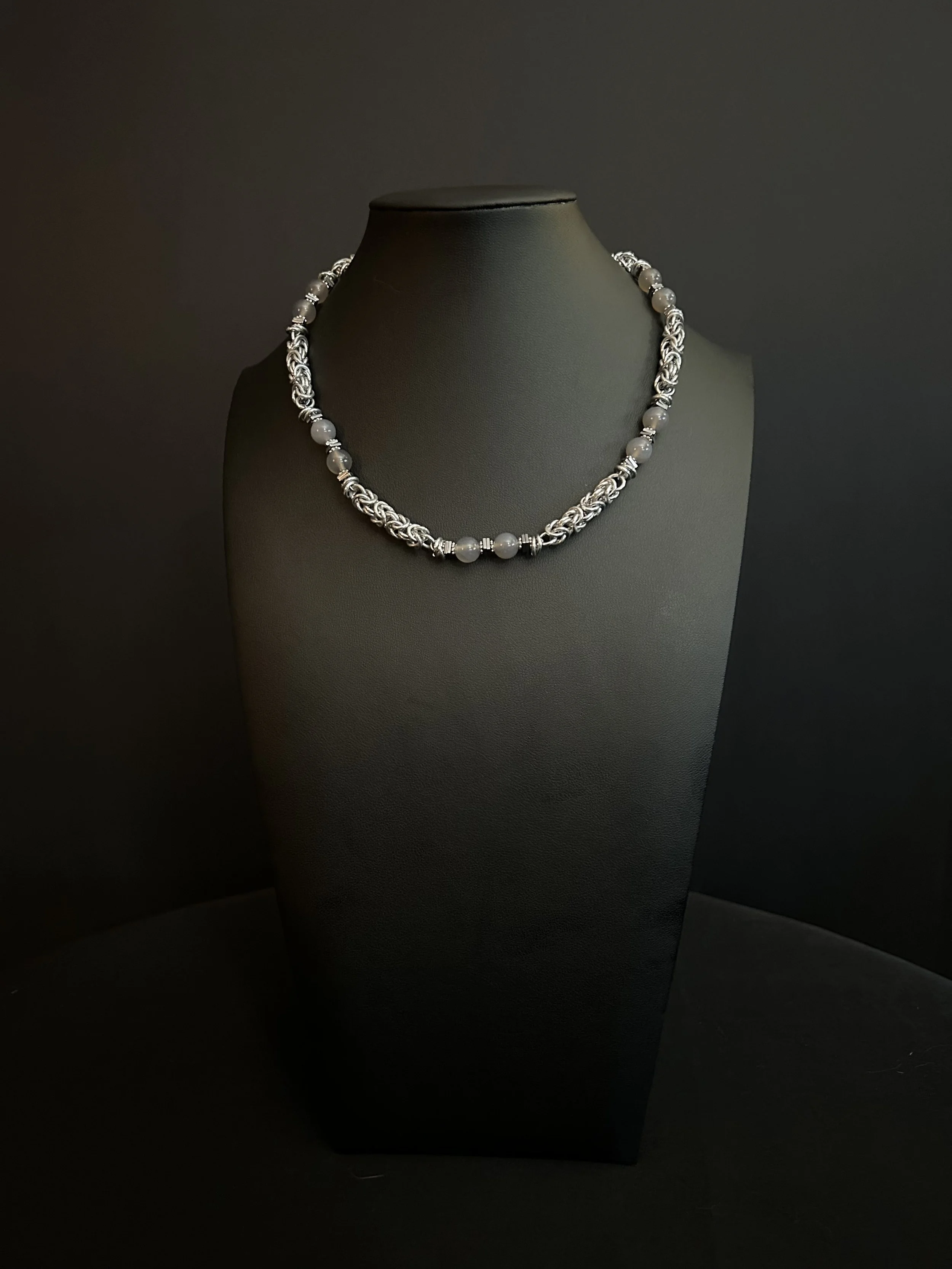 Moonlit Harbor necklace displayed on a black jewelry bust, featuring gray chalcedony beads, hematite spacers, and handwoven aluminum chainmaille.
