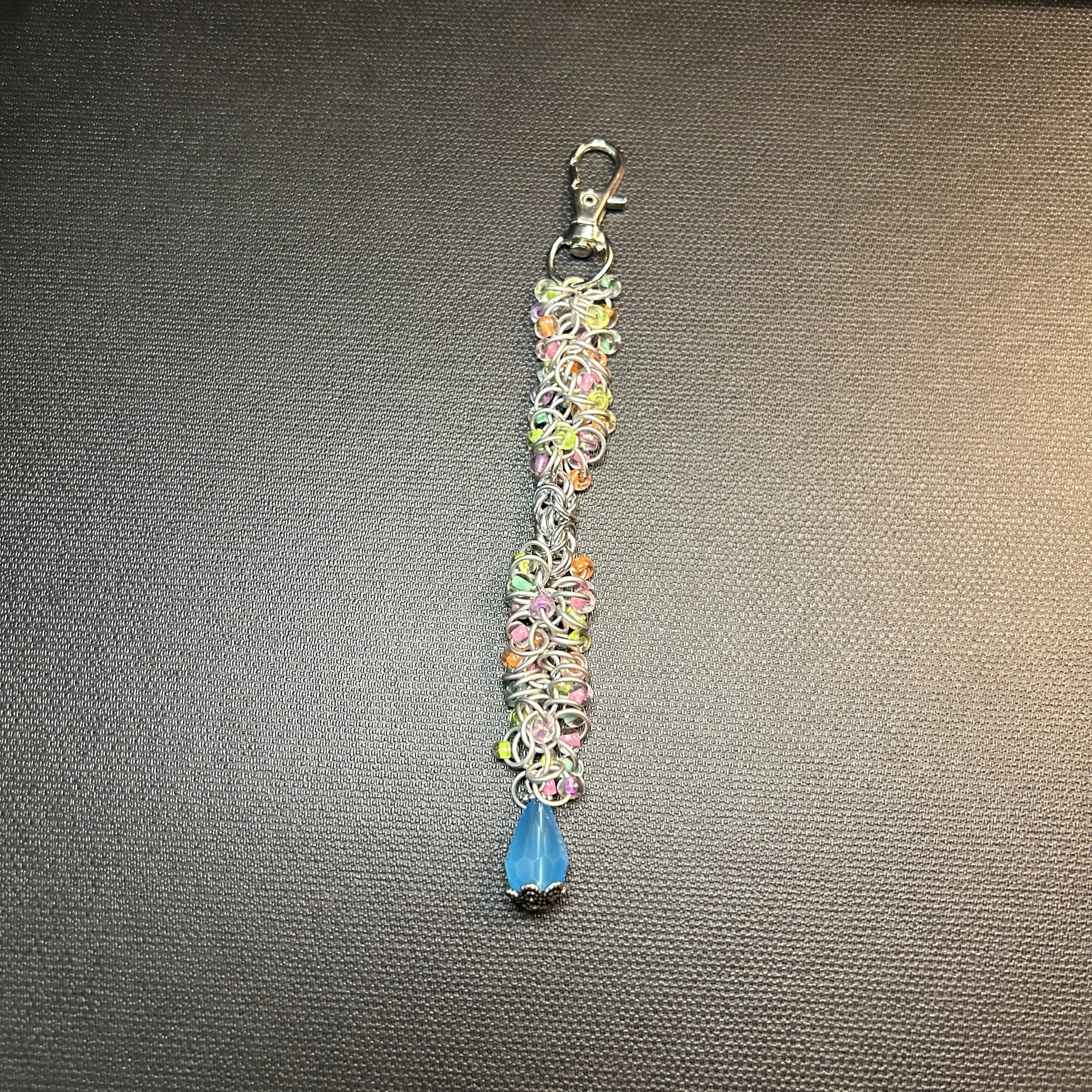 Silver shaggy loop and Byzantine purse charm shown flat on a dark surface with pastel bead accents and a blue drop.