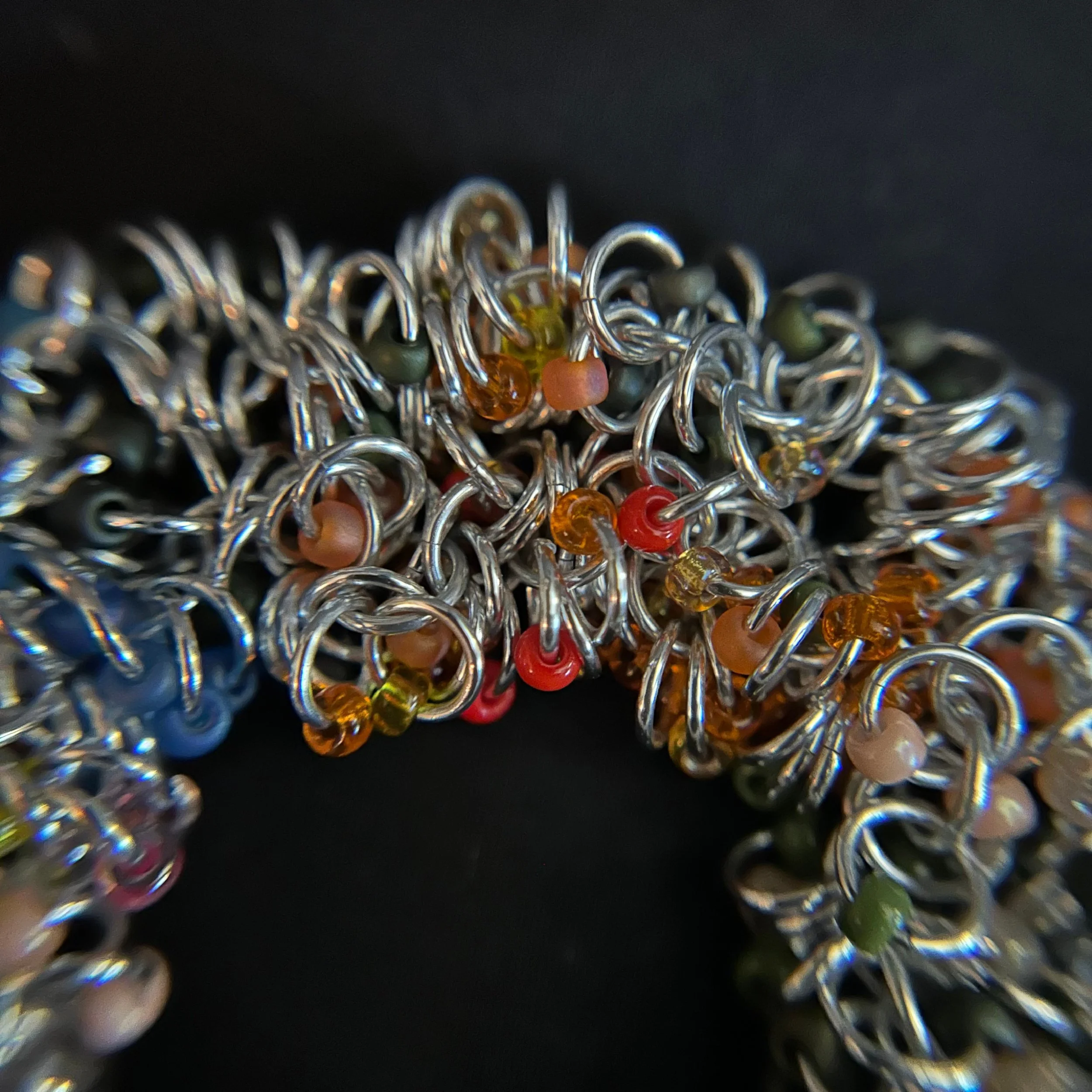 “Detail of the chainmaille rings with red, gold, green, and orange beads.”“Detail of the chainmaille rings with red, gold, green, and orange beads.”