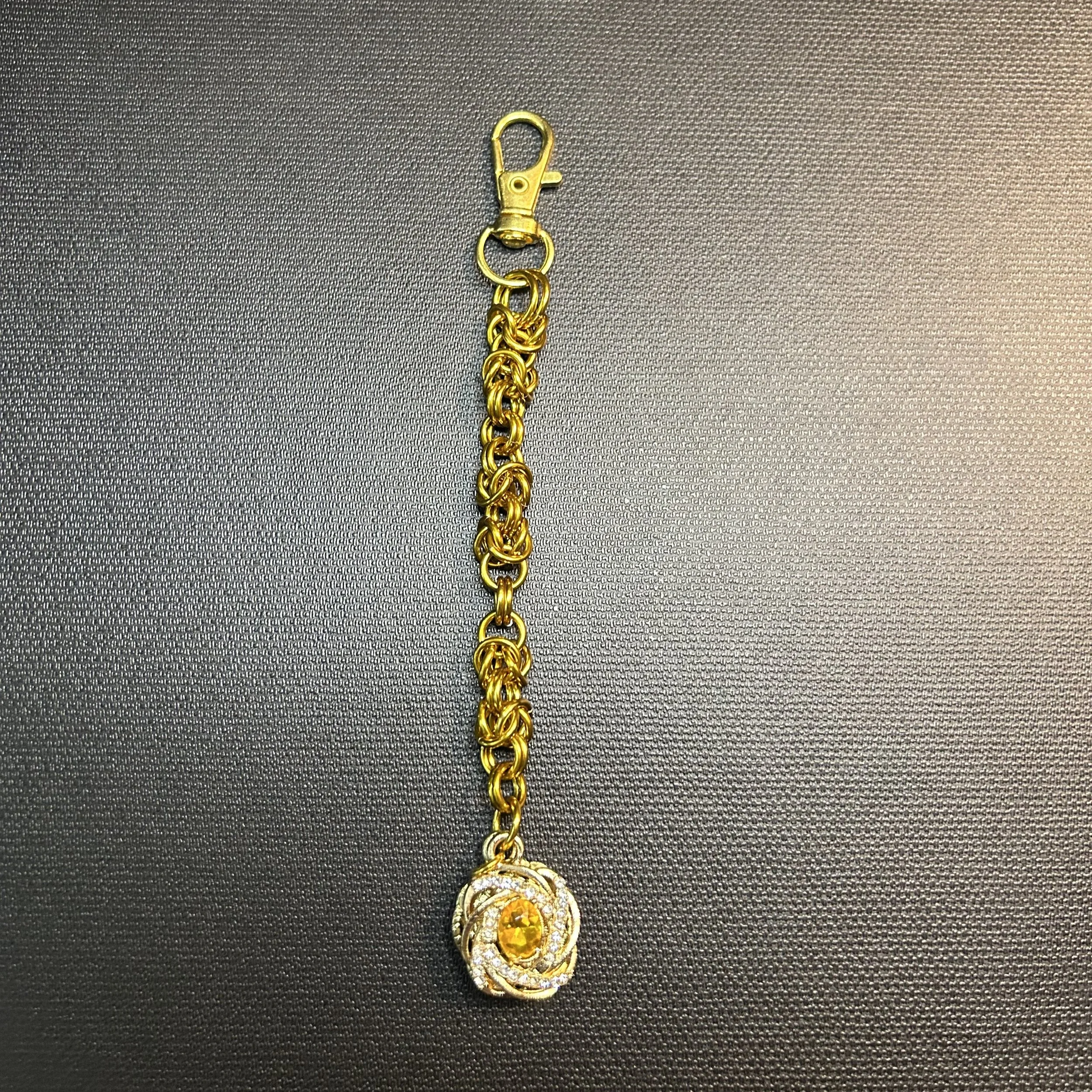Gold‑tone Byzantine chainmaille purse charm shown flat on a dark textured surface with a clear pave and gold crystal focal and swivel clasp.