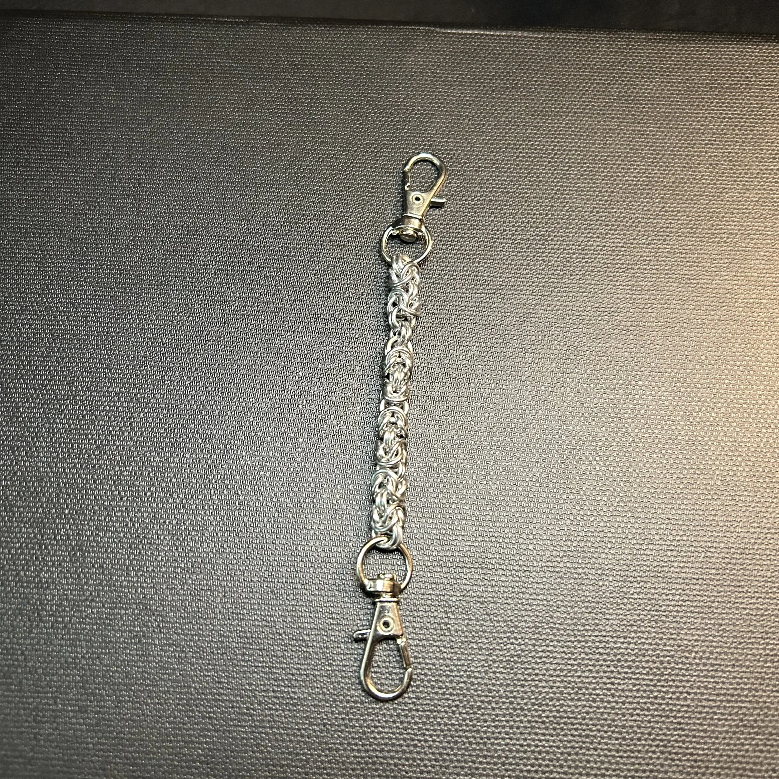 Silver chainmaille bag chain with dual swivel clasps shown flat on a dark textured surface.