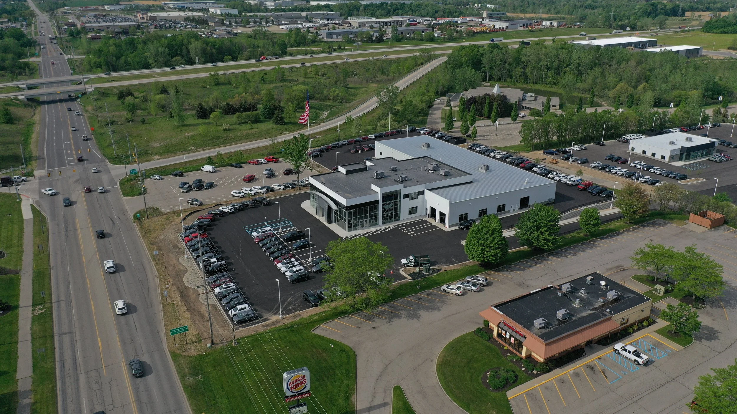 Aerial view of a commercial area including a car dealership, and several office or retail buildings, with green trees and roads surrounding the area.