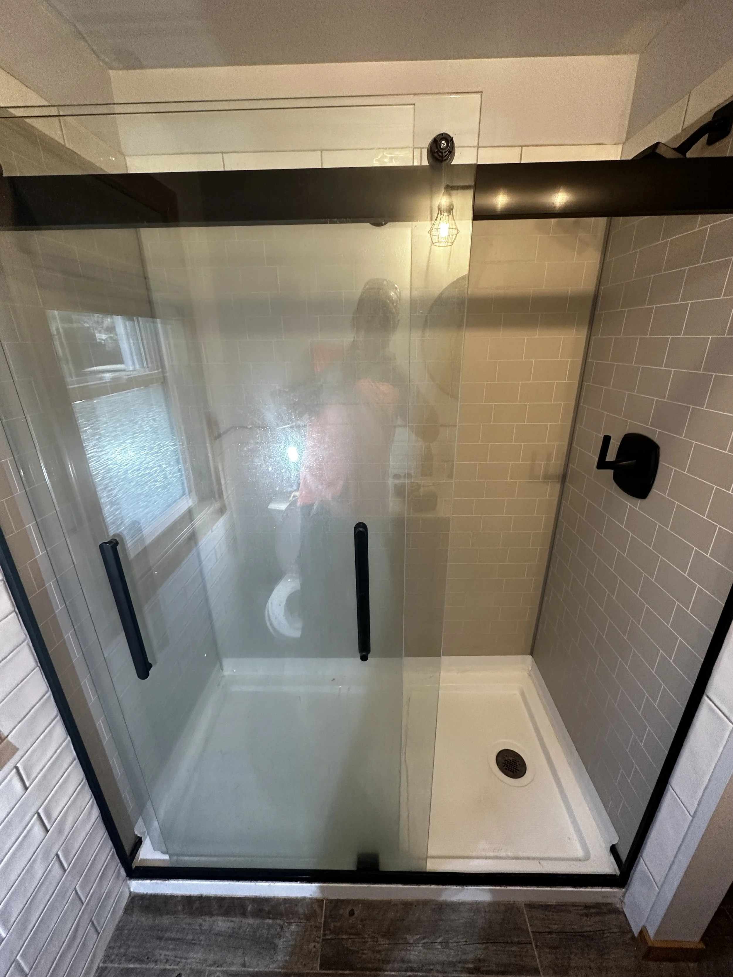 A glass shower enclosure with black handles, beige tiled walls, a small white shower pan, and a window visible in the reflection.